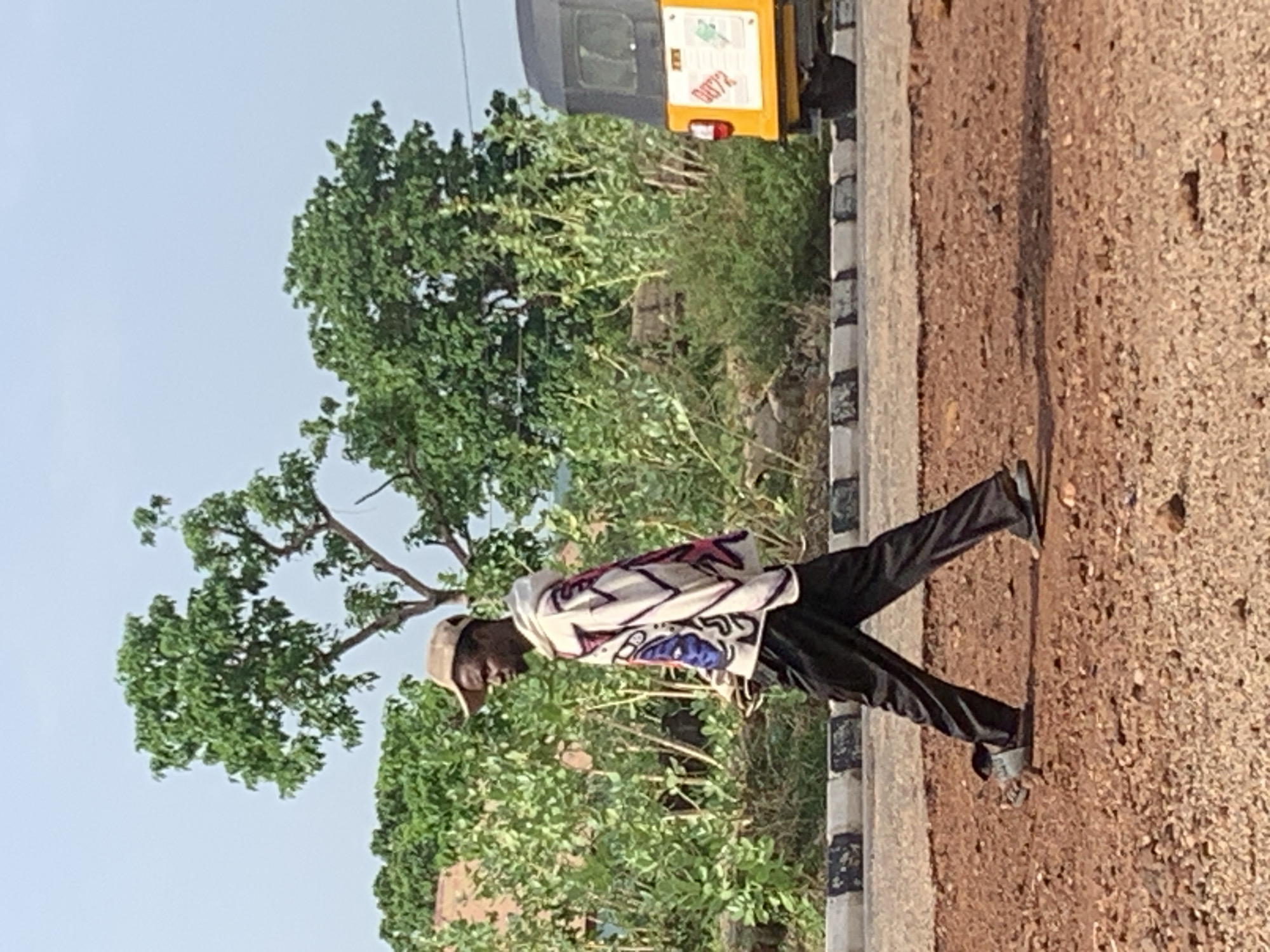 In Adankolo, Nigeria, an individual is seen walking on the right side of a paved road. The person is wearing black pants, a light-colored shirt with a graphic design, a beige cap, and dark shoes. The road is bordered by a curb painted with alternating black and white stripes. To the left of the road and individual, lush green vegetation fills the scene, partially obscuring a yellow three-wheeled vehicle (tuk-tuk) with a registration number (0072) on its rear. The ground to the right of the road is covered in dirt and small rocks. The sky is visible at the top of the frame.