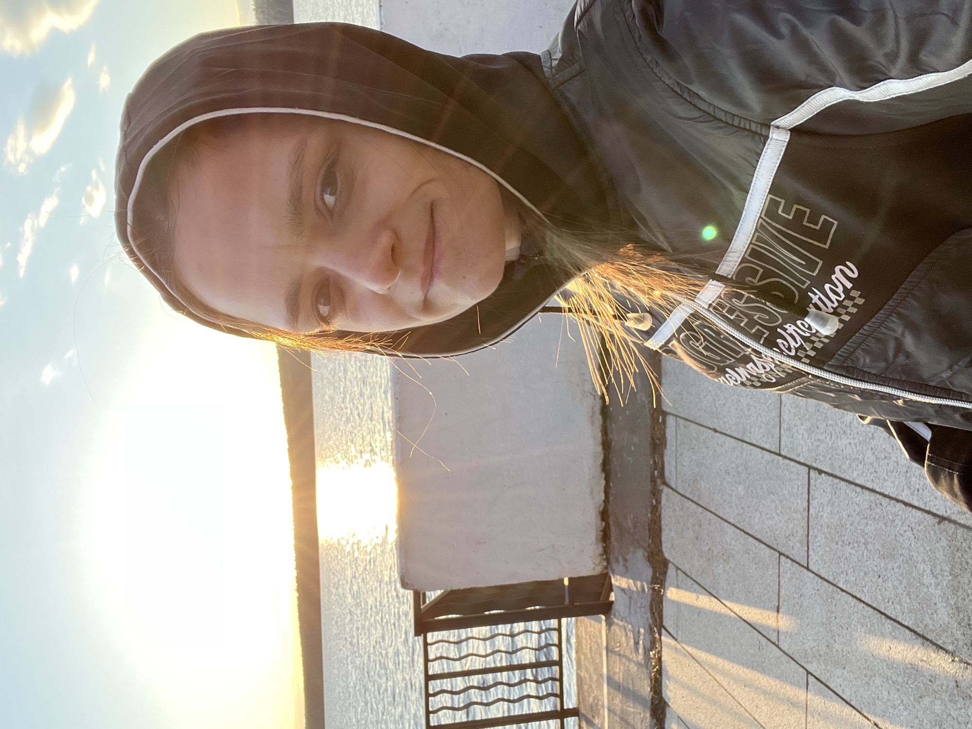 The image captures an outdoor portrait, likely a selfie, of a young person in Chaikovsky, Russia, taken during sunrise or sunset. The subject, who appears to be female, is positioned prominently in the foreground, looking directly at the camera with a gentle, relaxed smile. She is wearing a black hooded jacket with white stripes or piping on the sleeves and front. Her light-colored hair glows with a golden light, a result of the strong backlight.
The scene is set alongside a large body of water, likely the Kama Reservoir, which occupies the left and central background. The sun is low on the horizon, creating an intense, warm glare and a shimmering reflection across the water's surface. On the right side of the frame, there's a paved area, possibly a promenade or boardwalk, with light-colored rectangular tiles. A white, low wall or structure and a dark metal railing are also visible, bordering the water. The sky is clear, with a few faint clouds in the distance.
The dominant feature is the warm, golden light of the low sun, which bathes the entire scene in a soft, bright glow, highlighting the subject's hair and reflecting brilliantly off the water. The person's expression conveys a sense of peaceful enjoyment. Visible text on the jacket includes parts of words like "STRE" and "retro dition," accompanied by a small checkered pattern.