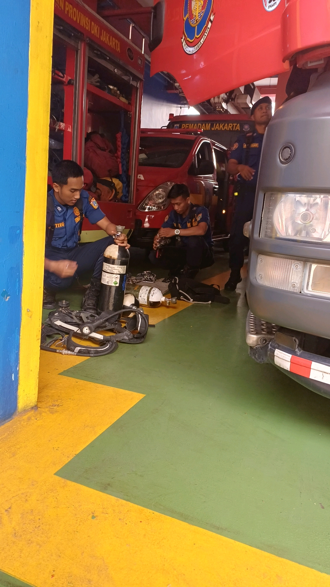 Two firefighters, wearing navy blue uniforms with orange trim and black boots, are working on breathing apparatuses inside a fire station. The firefighter on the left is kneeling and holding a large black oxygen cylinder, preparing to attach it to a breathing apparatus frame lying on the green and yellow painted floor. The firefighter on the right is also kneeling and appears to be working on a similar breathing apparatus. A red fire truck with "PEMADAM JAKARTA" (Jakarta Fire Department) written on it is visible in the background, along with another red truck and a grey utility vehicle. The environment is a well-lit interior space with a painted floor marking a clear division between yellow and green sections. The location is identified as a fire station in South Jakarta, Indonesia.