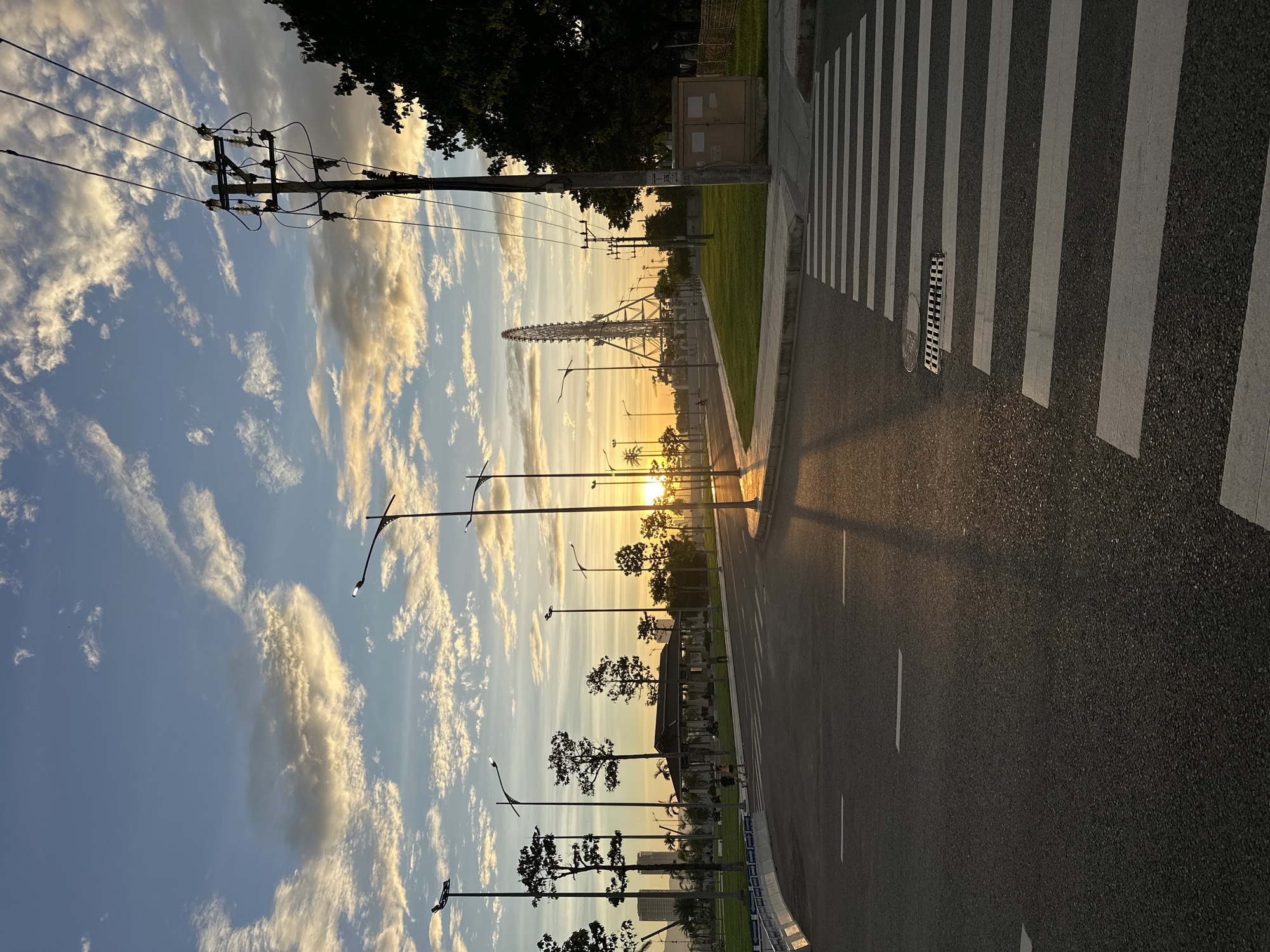 The image depicts a wide asphalt road with white lane markings and a pedestrian crosswalk in the foreground. The sun is low on the horizon, casting long shadows and creating a warm, golden glow. The sky is a blend of blue and white, with scattered clouds illuminated by the setting sun.
In the background, a Ferris wheel stands tall against the sky, indicating the location is likely an amusement park or recreational area. Trees line the sides of the road, and streetlights are visible, some illuminated. Several people are seen walking on a paved pathway to the left of the road. A power pole with numerous wires stands to the right, along with a small electrical box. The overall impression is a serene urban landscape at sunset, possibly in the city of Đà Nẵng, Vietnam, given the presence of a Ferris wheel which is a prominent landmark in the coastal city.