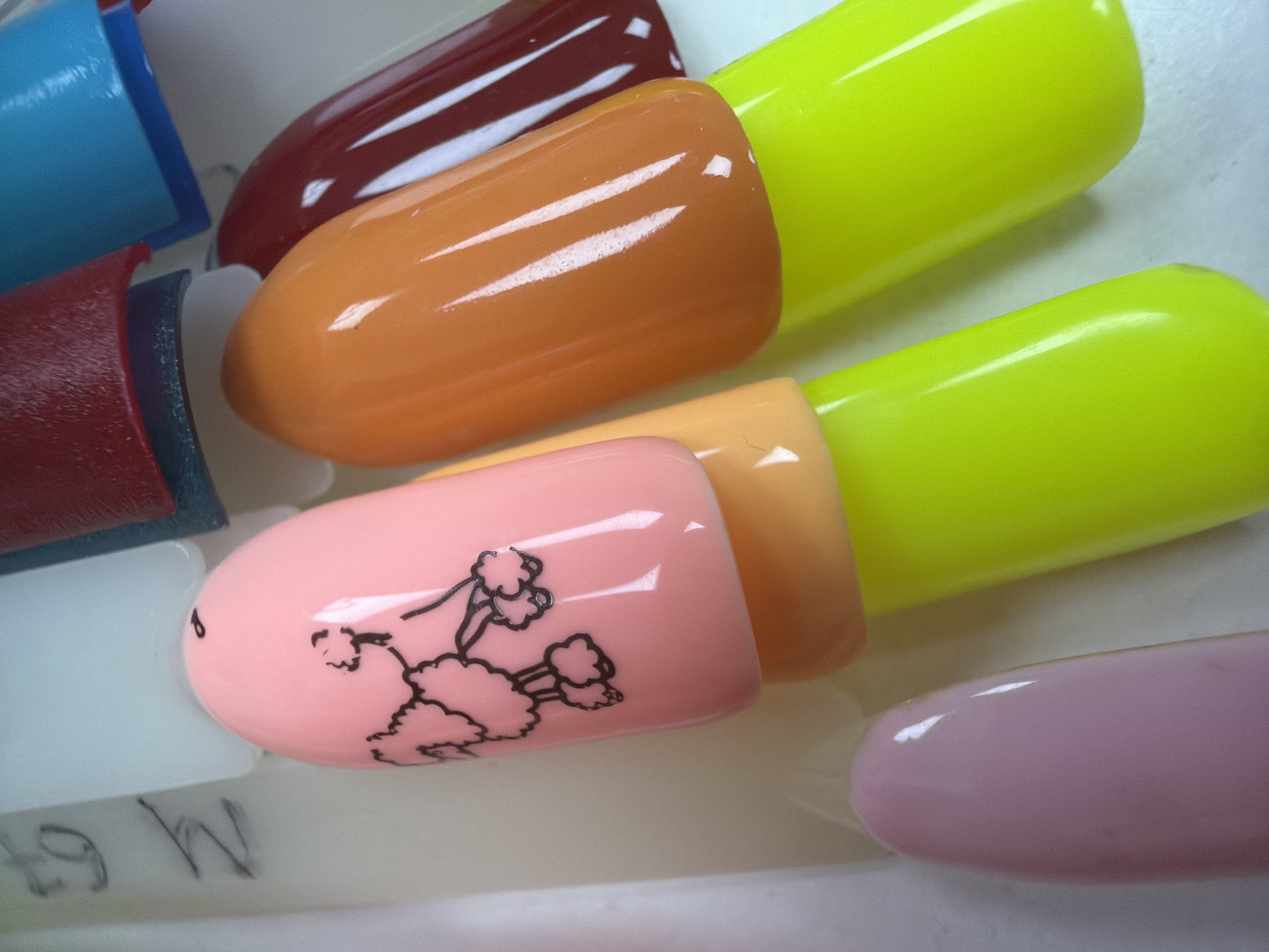The image is a close-up, overhead shot of several nail polish swatches on a white surface, with some black lettering visible in the background. The swatches are arranged in a diagonal pattern. From left to right, the visible swatches are: a pale pink oval shape, a pink oval shape with a black poodle design, a light orange oval shape, and a dark red oval shape. Below the pink swatch with the poodle design is a bright lime green oval shape, and to its right is another bright lime green oval shape. The lettering in the background appears to be a handwritten code, possibly related to the colors or types of polish. The lighting creates reflections on the glossy surfaces of the nail polish. The scene is indoors, on a flat surface, with no indication of any specific outdoor location or city.