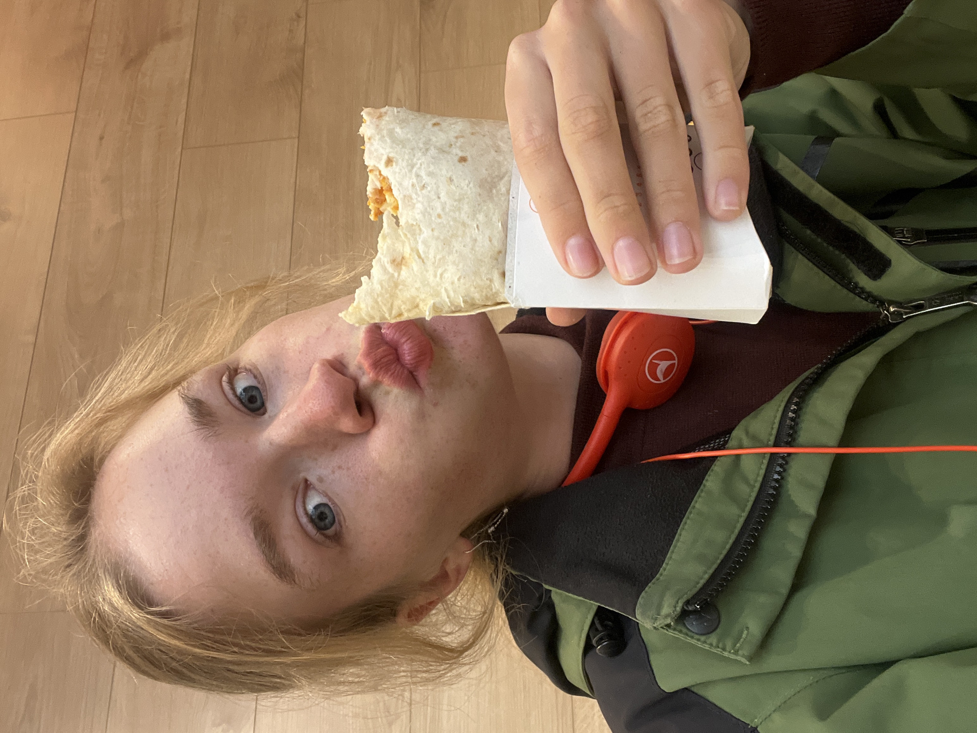 A young, fair-skinned person with light blonde hair and blue eyes is holding a partially eaten burrito in their right hand. They are positioned in front of a wooden floor. The person is wearing a dark burgundy shirt underneath an olive green jacket. Orange headphones are visible near their collar. The person has their lips pursed as if posing for a selfie. The location is unknown and there is no information to suggest this photo was taken in Budva, Montenegro.