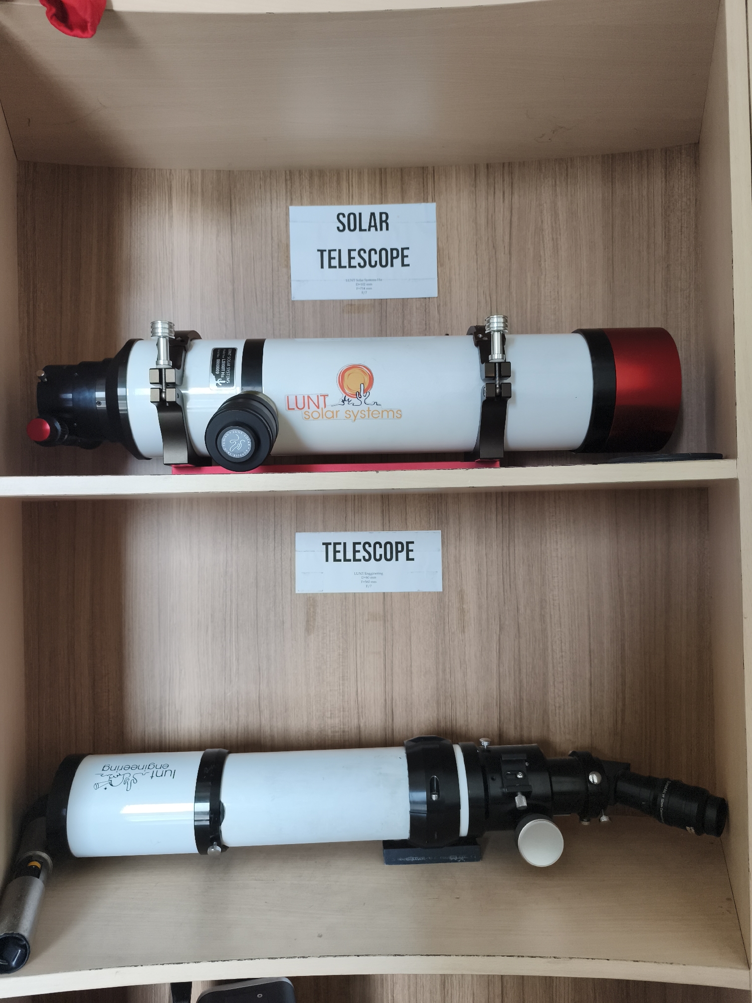 The image shows two Lunt telescopes displayed on wooden shelves. The upper shelf holds a white Lunt solar telescope with a red end cap and a "LUNT solar systems" logo. Above it, a sign reads "SOLAR TELESCOPE". The lower shelf contains another white Lunt telescope, oriented horizontally, with black accents and a more complex eyepiece assembly. Above this telescope, a sign reads "TELESCOPE". The background is a light-colored wood grain pattern. The overall setting appears to be an indoor display or storage area. There is no indication of the city of Semarang, Indonesia in the image.