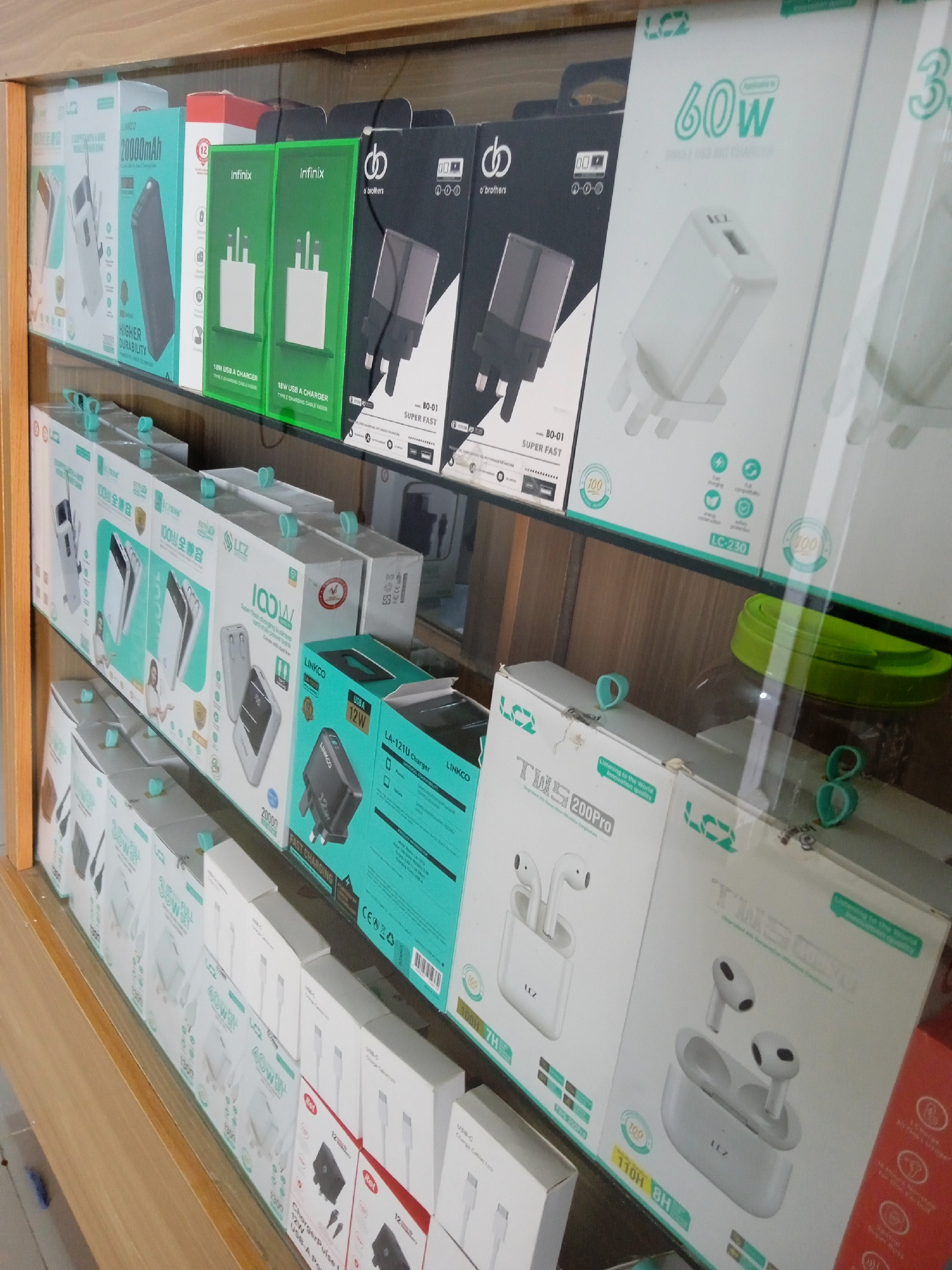 The image shows a display case filled with various electronic accessories, likely in a retail setting. The items include power banks, chargers, and wireless earbuds, from brands such as Infinix, o'brothers, LINKCO, and LCZ. There are different wattage chargers ranging from 12W to 100W. The products are neatly arranged on the shelves. The location, Jos, Nigeria is not directly evident in the image.