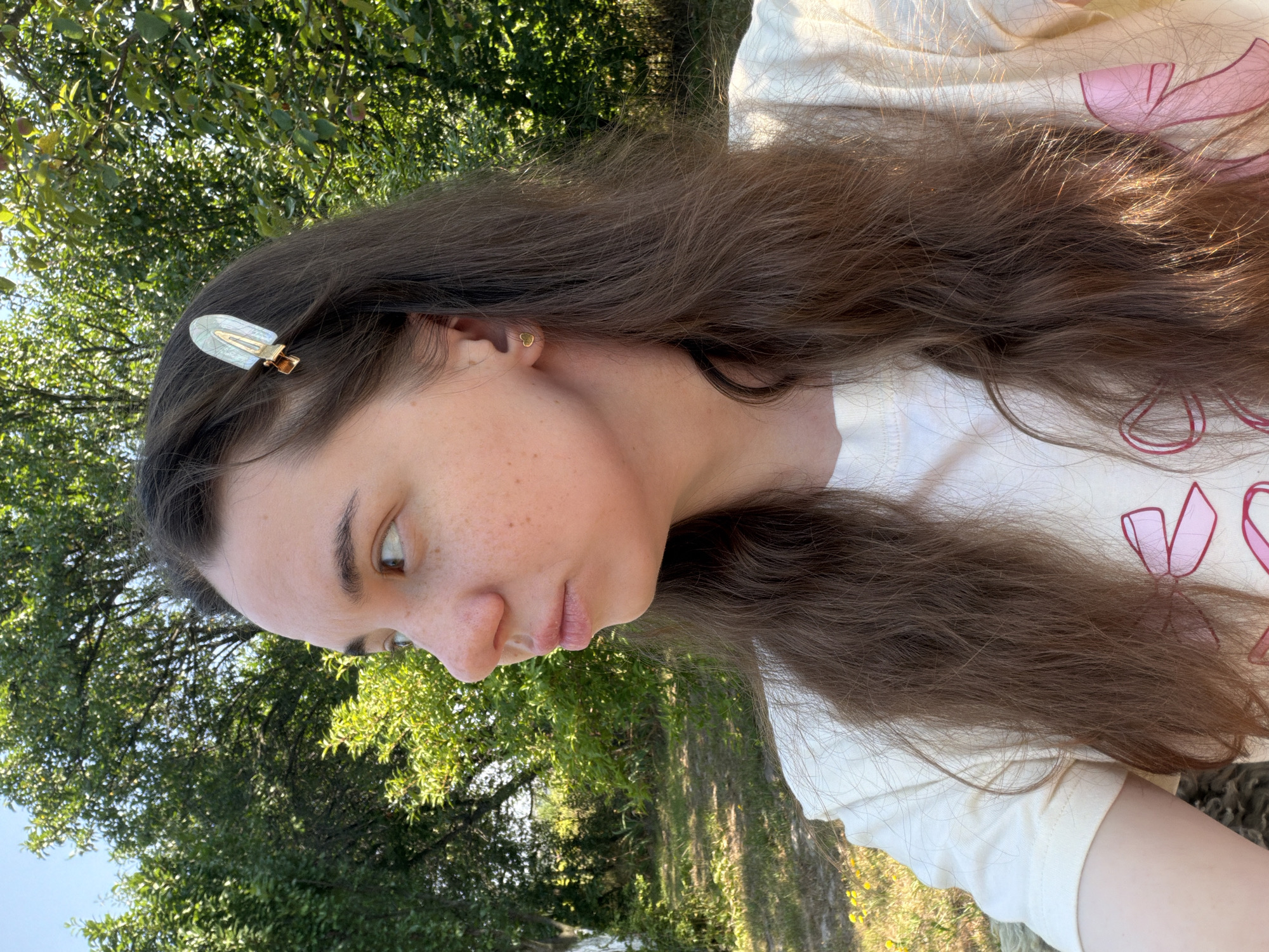 A young woman with long brown hair and a pale complexion is captured from the chest up, turned to her left, against a backdrop of lush green trees. She is wearing a light-colored t-shirt adorned with pink bow graphics. A decorative hair clip, oval-shaped with a mother-of-pearl-like design and a gold clasp, is fastened to the side of her head. A small gold heart earring is visible in her left ear. Her face displays a pensive expression, with her lips slightly pursed and her gaze directed off-camera. The lighting suggests an outdoor setting, likely during the daytime, given the bright sunlight illuminating her hair and casting subtle shadows. The background consists of foliage, indicating a natural, possibly rural or park-like environment, consistent with the location of Mali Kosharyshcha, Ukraine.