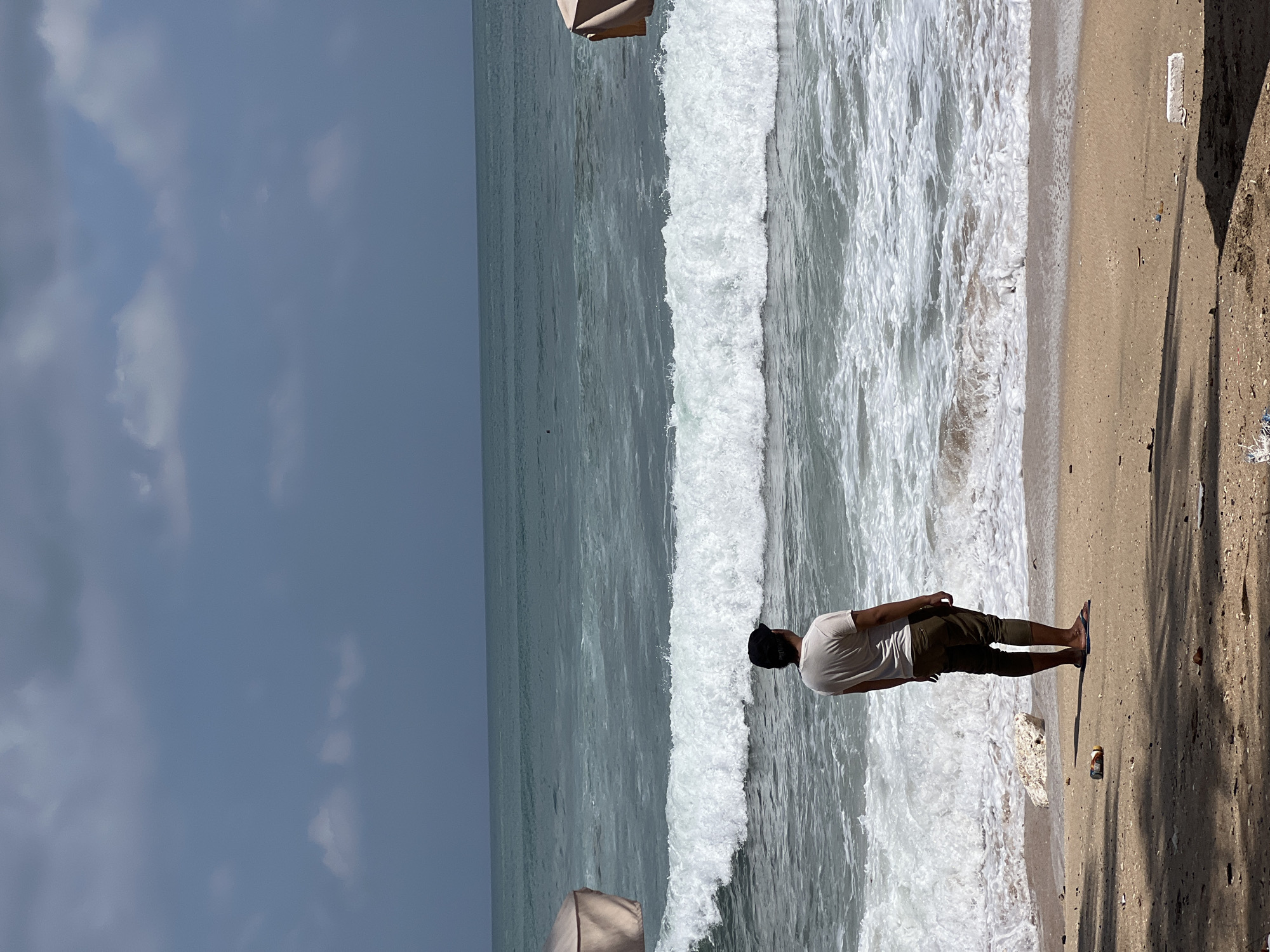 A lone individual stands on the sandy shore of Kuta Beach, Indonesia, facing the turbulent ocean. The sky above is a muted blue, partially obscured by scattered clouds. The sea is a dynamic expanse of turquoise and white, with a prominent wave breaking near the shore. The beach is a mix of sand and debris, with a discarded bottle visible in the foreground. Two beach umbrellas, partially visible on the left and right edges of the frame, suggest a recreational area. The individual, wearing a light-colored t-shirt, dark pants, and a dark cap, appears to be contemplating the vastness of the ocean. The overall scene conveys a sense of solitude and natural beauty, juxtaposed with the presence of human-made waste.