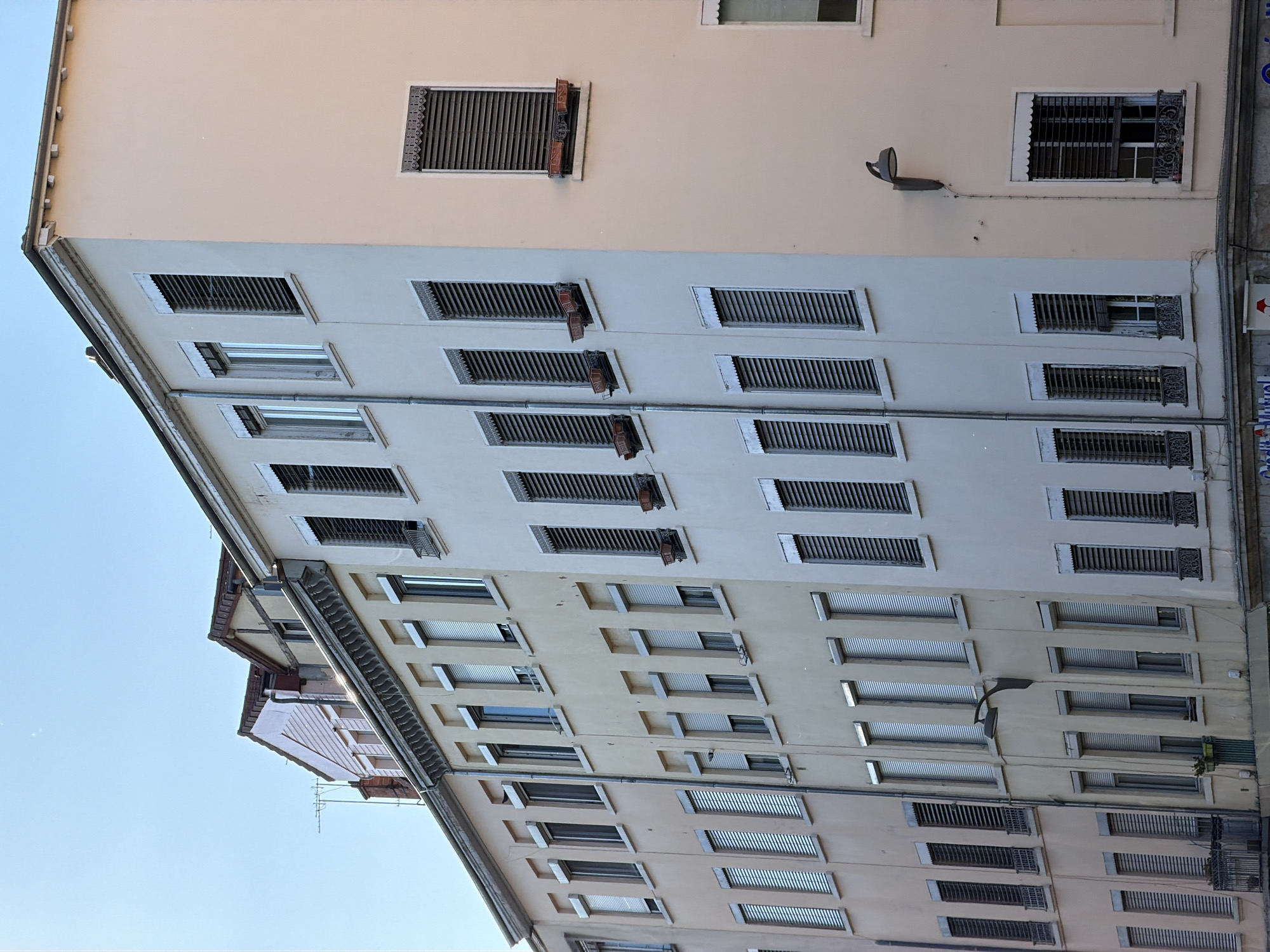 The media file displays a close-up, low-angle view of the facades of several buildings in Lyon, France, under a clear sky. The buildings are multi-story structures with a variety of windows, many of which are covered by adjustable louvers, also known as "jalousies." The architecture suggests an urban setting, likely a street lined with apartments or commercial spaces.
The sunlight indicates it is daytime, and the clear sky suggests fair weather. The buildings have a light-colored stucco or painted exterior, with the lower sections appearing slightly darker or a different shade than the upper stories. Details visible include window frames, some with decorative ironwork, and external conduits or piping running along the walls. On the upper part of the central building, a modern-style outdoor lamp is mounted on the wall.
The scene is static, with no people or overt activity captured. The focus is purely on the architectural elements and the play of light and shadow on the building surfaces. The orientation of the image is rotated 90 degrees counter-clockwise, presenting the buildings as if viewed from the side and angled upwards. No legible text is visible in the image.