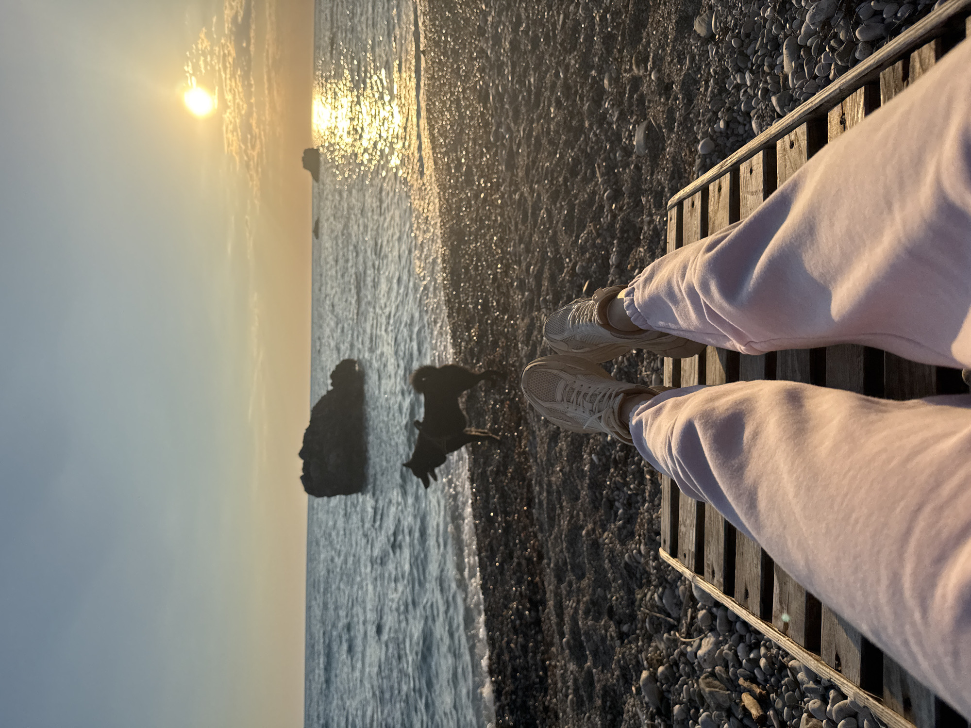 A vertical medium shot shows a person sitting on a wooden boardwalk at a beach in Bar, Montenegro, facing the sea at sunset. The scene captures the subject from the knees down, wearing light-colored sweatpants and sneakers. The boardwalk is situated at the edge of a pebble beach that meets the water. The sea is calm, with a few rock formations visible in the distance. The sun is setting over the water, creating a bright glow and reflections on the surface of the sea. A dog, appears to be moving on the beach, between the boardwalk and the water. The overall lighting is warm due to the sunset. The pebbles on the beach are dark and wet.