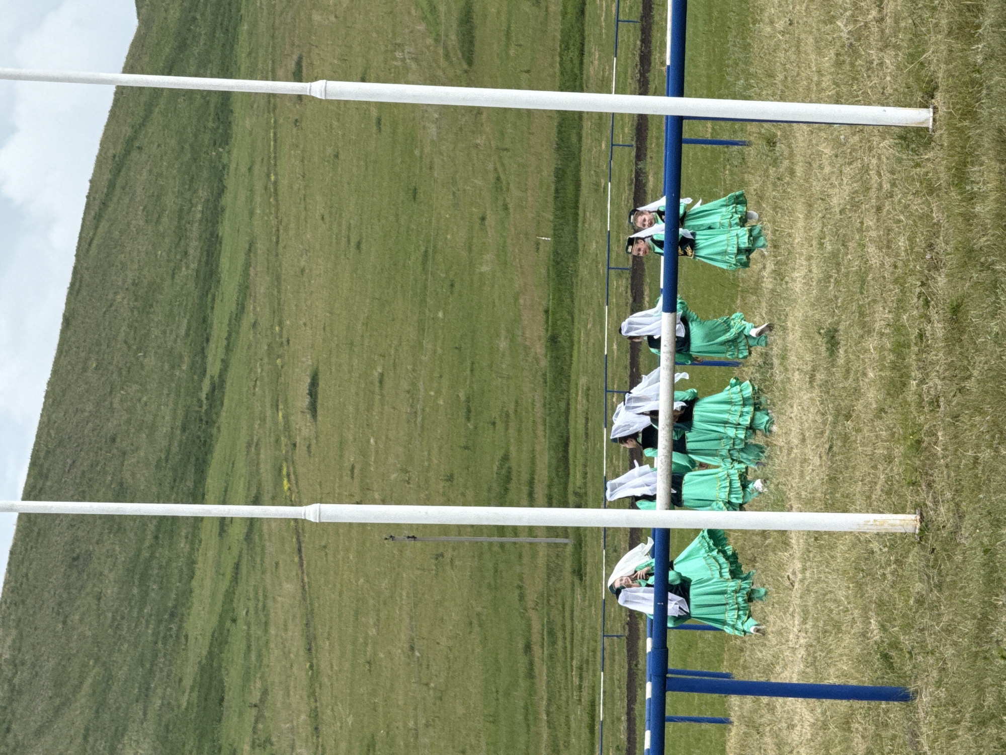The image depicts a group of girls in traditional green dresses and white headscarves performing outdoors on a grassy field. The background features a rolling green hill under a cloudy sky. In the foreground, a blue and white railing runs horizontally across the scene, with two white poles positioned in front of it. The girls are positioned behind the railing, some facing the camera and others with their backs turned. One girl appears to be holding a phone. The setting suggests an outdoor performance or cultural event.