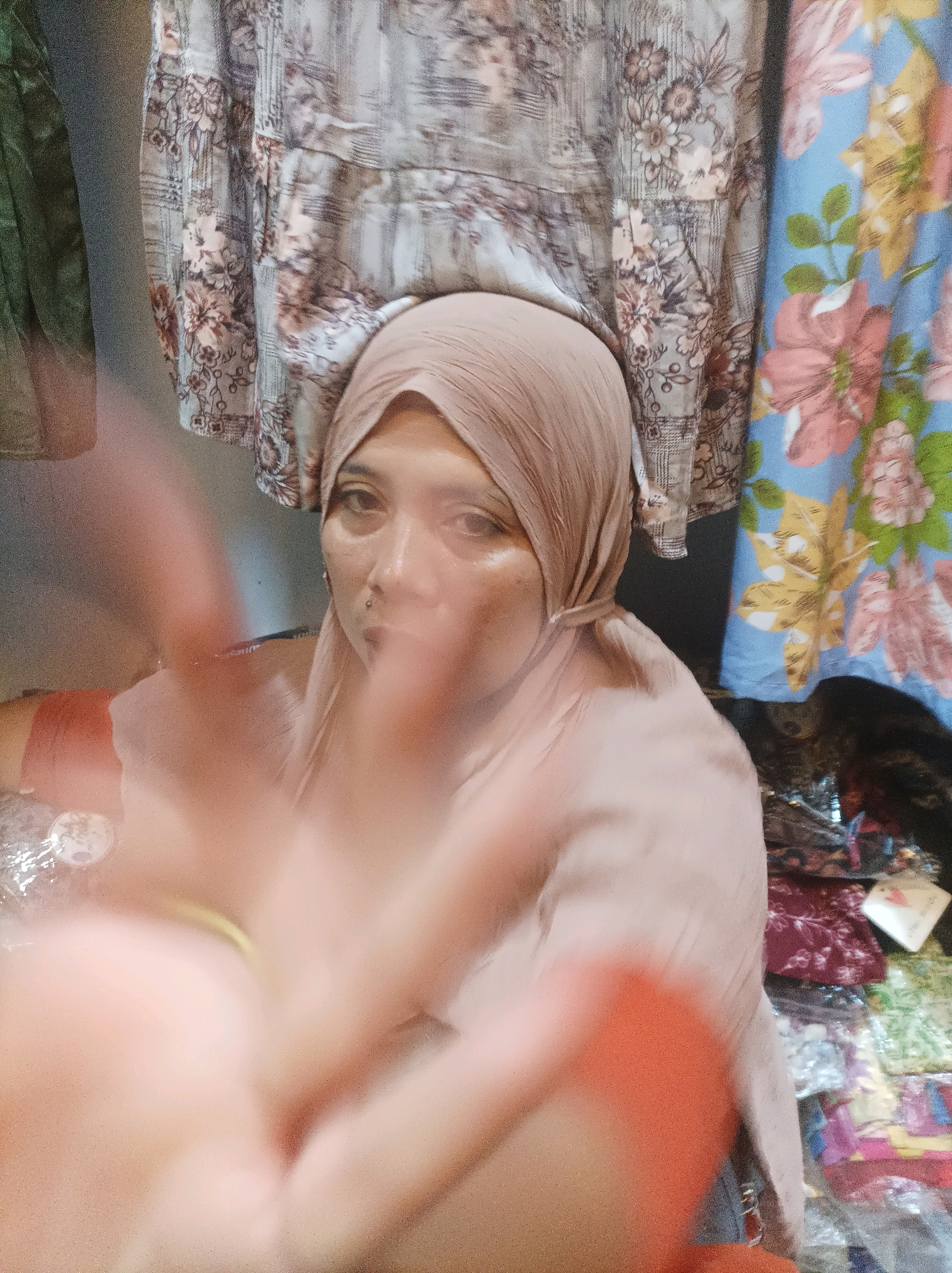 The image shows a close-up of a woman wearing a light brown hijab and a matching headscarf. Her face is slightly blurred, and she has dark eyes, visible eyebrows, and a pierced lip. She appears to be in an indoor setting with patterned fabrics hanging in the background. To the left, a green fabric is visible, and to the right, a blue fabric with large floral patterns is present. In front of the woman, a hand is obscuring part of the image, with fingers extended in what appears to be a peace sign. There are also piles of fabric or clothing in the lower right portion of the image. The scene is consistent with a modest clothing store or a personal collection of fabrics. There is no specific indication of the location within Sumuragung, Indonesia.