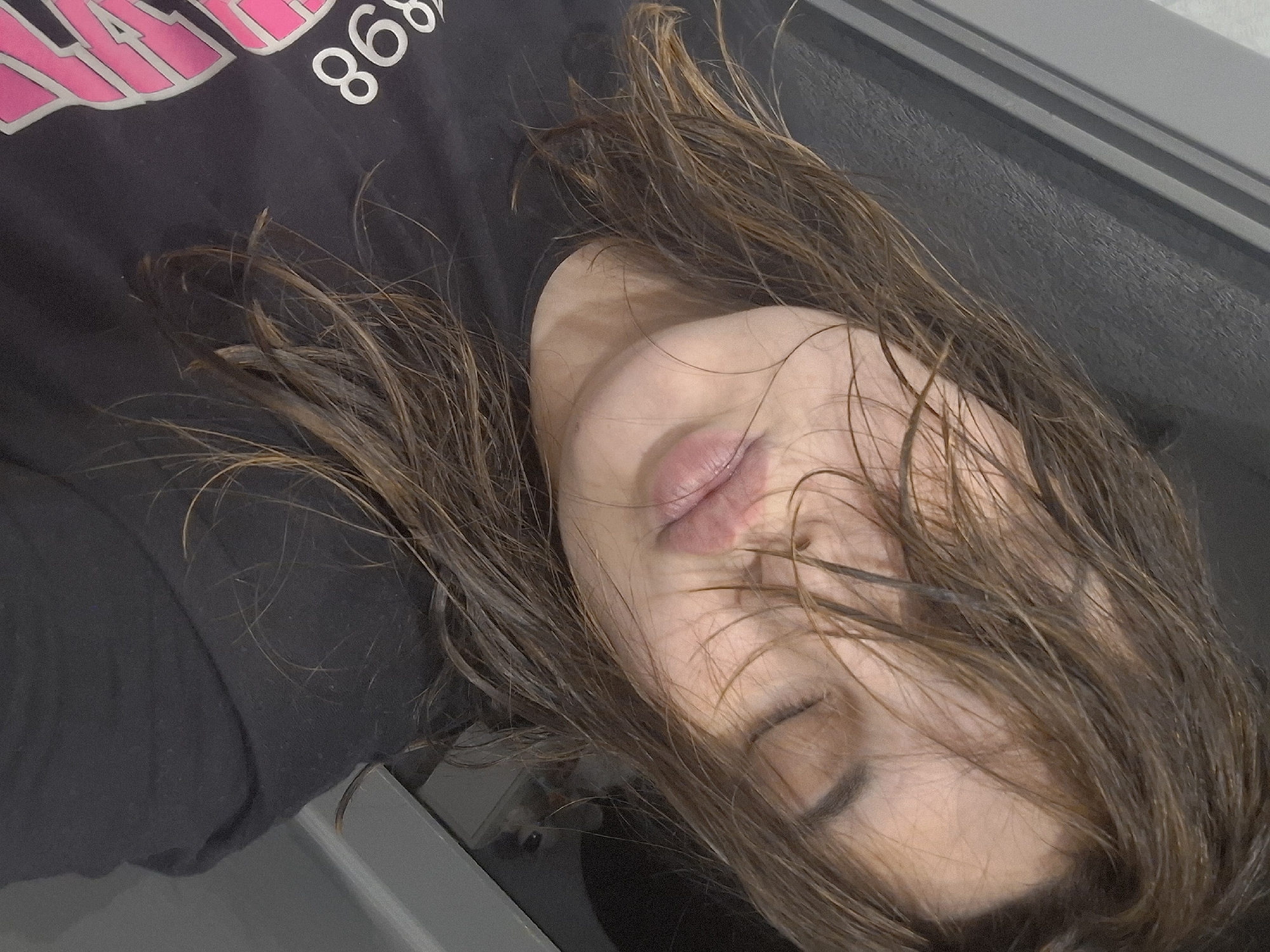 The image captures a close-up view of a young adult female, positioned diagonally with her head tilted to the viewer's right. Her eyes are closed, and her lips are pursed into a 'duck face' or 'kiss' expression. Her medium brown hair appears wet or disheveled, with strands falling across her face, obscuring parts of her forehead and right eye. She has a fair complexion. The individual is wearing a dark, short-sleeved t-shirt, on which white numbers "898" are visible on the upper left chest area, accompanied by a partial pink design beneath them. The background consists of indistinct dark gray and black surfaces, indicative of an indoor setting, with no specific environmental features discernible to denote a particular location.