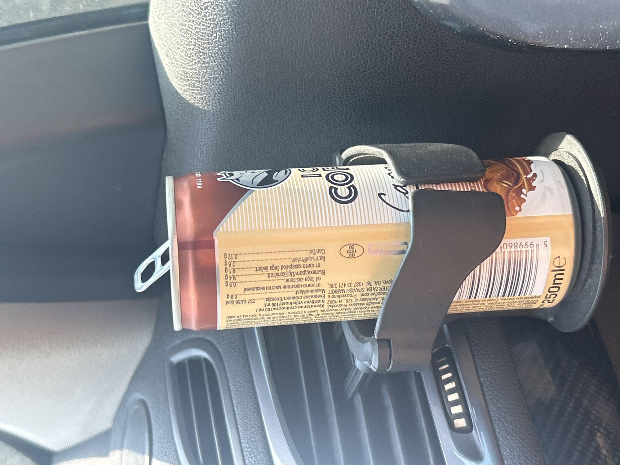 An opened can of "ICE COFFEE Caramel" from "HELL ENERGY" is secured in a car's cup holder. The can is mostly brown and gold with text printed in various languages, indicating it is a beverage. The cup holder is black and appears to be made of plastic, with a textured grip. The can is positioned in front of a dark grey, textured surface, likely the interior of a vehicle. Behind the can, a sliver of a car's air vent is visible, indicating the photo was taken inside a car. The lighting suggests daytime, with sunlight possibly filtering through the car's windows, creating shadows and highlights on the objects. The background is out of focus, but the overall environment appears to be the interior of a car, possibly parked in Plovdiv, Bulgaria, given the presence of Bulgarian text on the can.