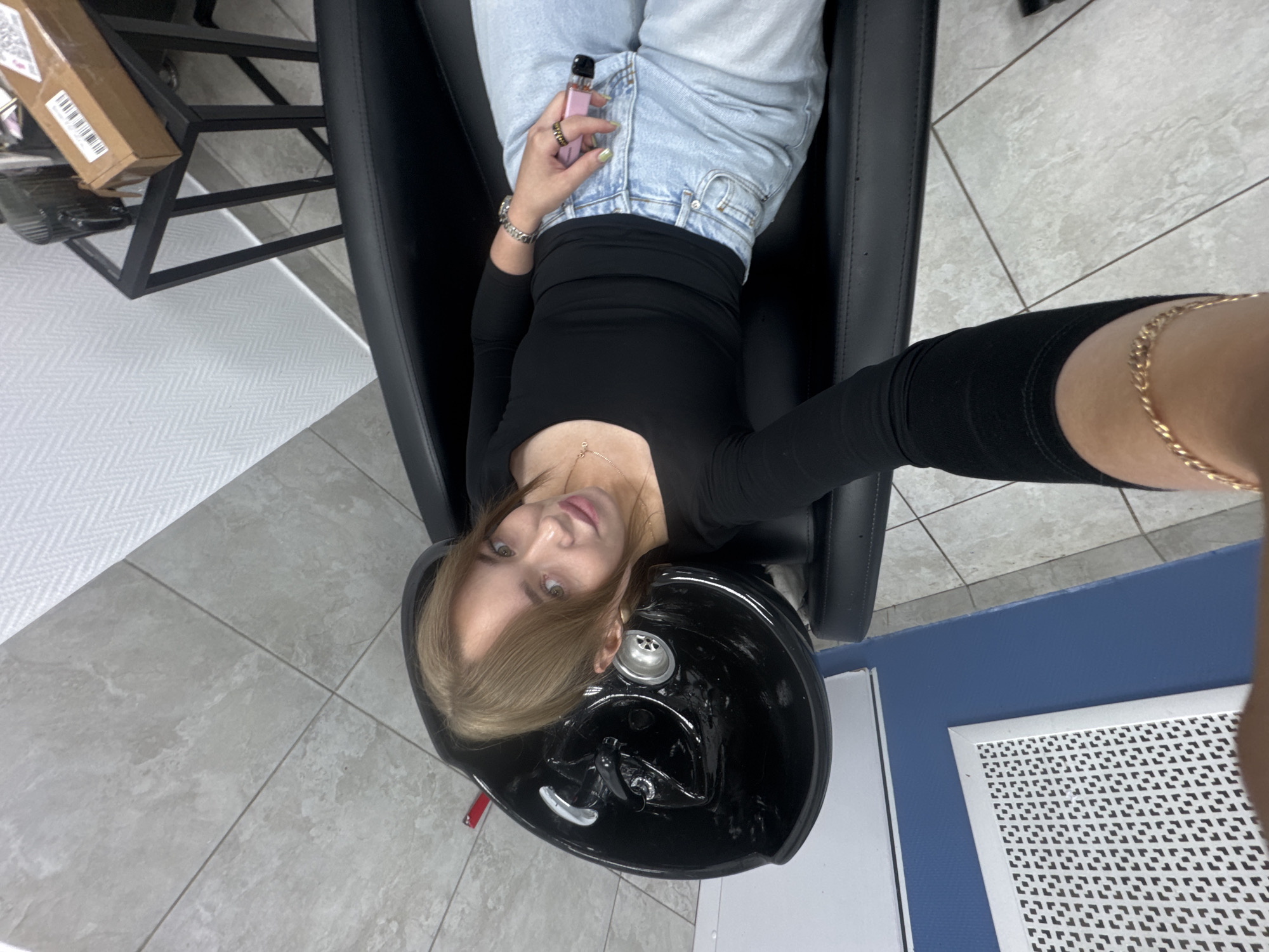 An overhead, wide shot captures a young woman lying in a black salon chair, positioned at a black shampoo bowl. She is looking upwards, toward the camera, with a slight smile. Her hair is styled in a way that frames her face, with bangs swept to one side and blonde highlights visible. She is wearing a black long-sleeved top and light blue jeans. Her right hand rests on her leg, holding a pink electronic cigarette. A silver bracelet adorns her left wrist. The background reveals a tiled floor and a glimpse of a white textured wall. The overall environment suggests a salon setting. The image does not provide specific geographical markers that would indicate its location in Chaikovsky, Russia.