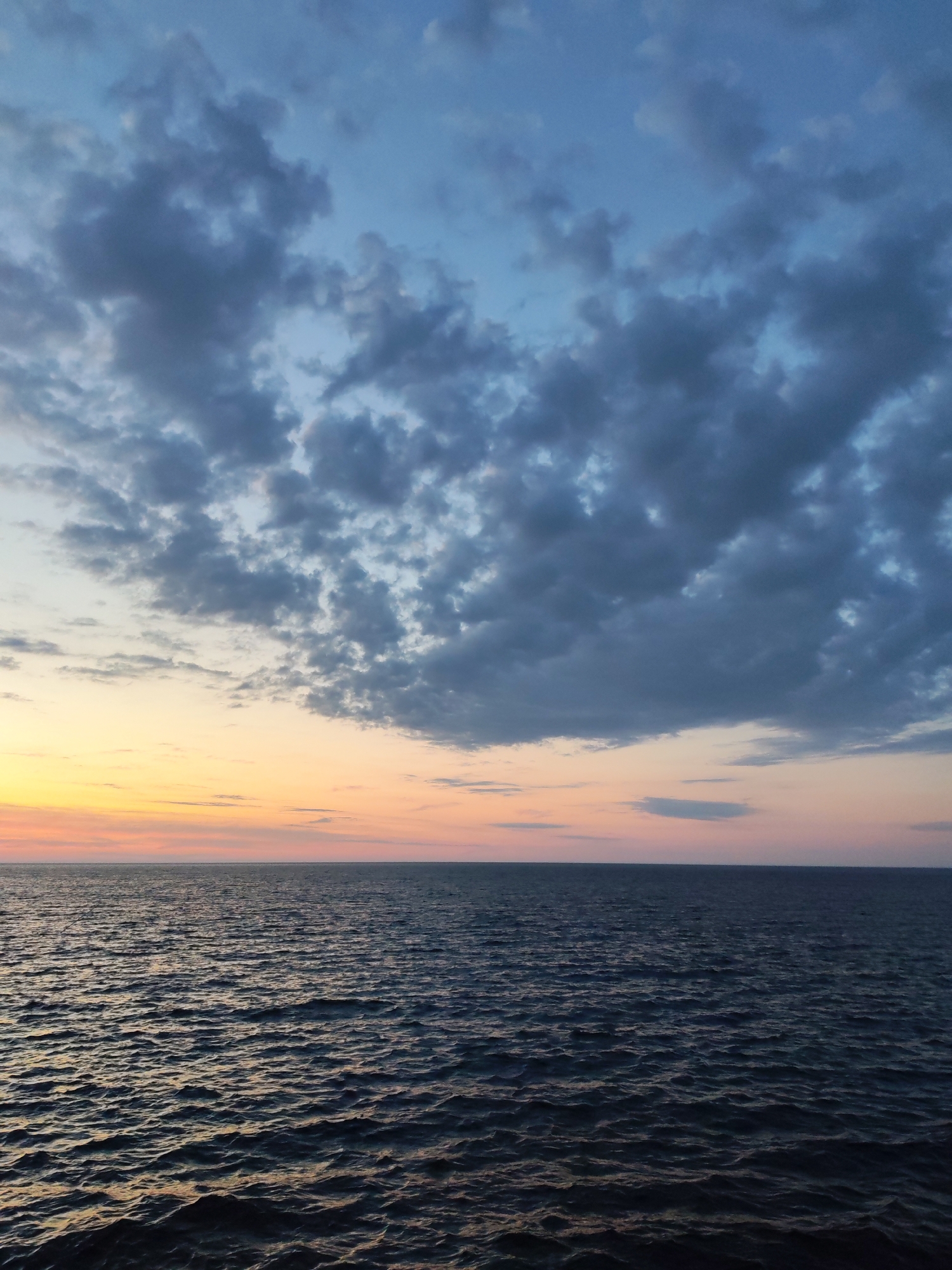 The image depicts a serene seascape at dusk. The sky is filled with a gradient of colors, transitioning from a soft peach and yellow near the horizon to a deep blue overhead. Scattered clouds, tinged with gray and white, drift across the sky. The ocean below is calm, with gentle ripples reflecting the muted colors of the sky. The horizon line is sharp and clearly defined, separating the sky from the water. There are no visible people, structures, or other man-made objects in the scene. The location is identified as Shcheleiki, Russia, implying this is a natural coastal landscape.
