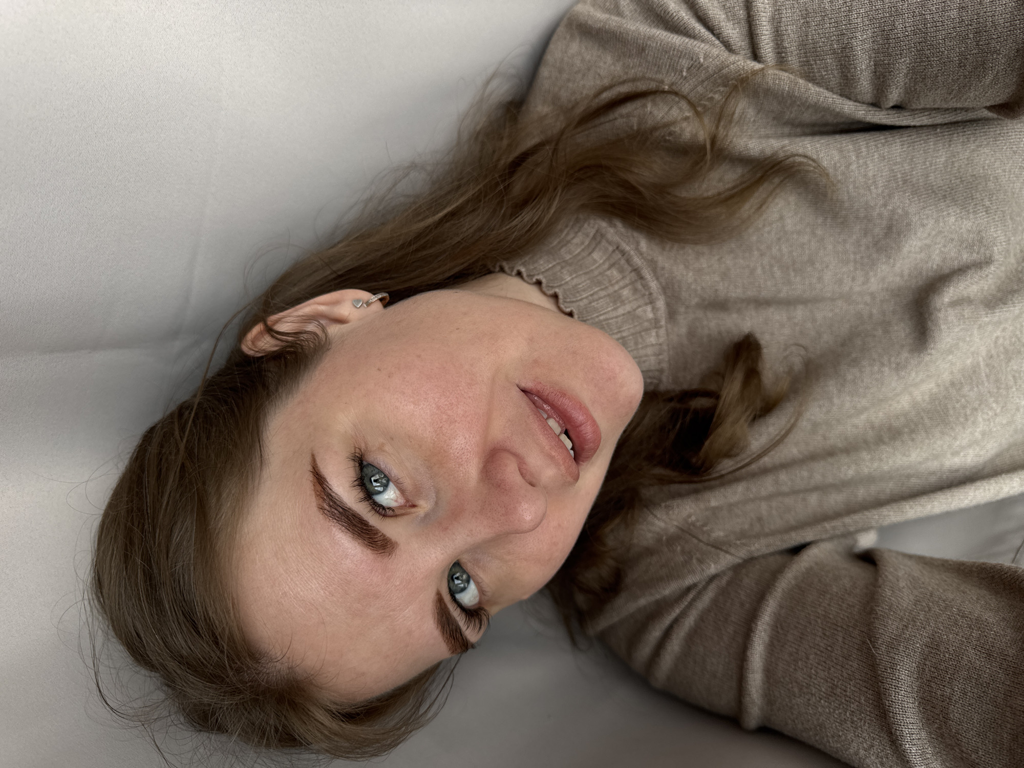 A woman with light brown hair and blue eyes is lying down, looking directly at the camera. She is wearing a light brown turtleneck sweater. Her face is close to the camera, and she has a silver earring on her left ear. The background appears to be a plain light gray or white surface. The location of the scene is unknown, but the prompt specifies Gornyi (Dvurechenskoe S/P), Russia.