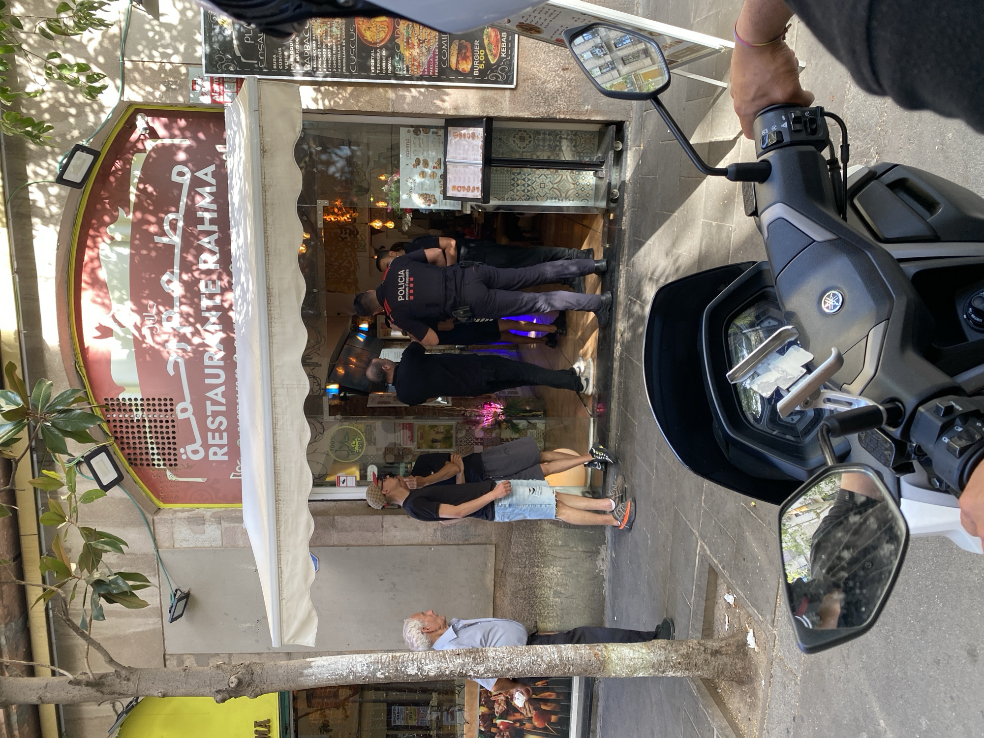 In Barcelona, Spain, outside a restaurant named "Restaurante Rahma" with Arabic lettering above, two police officers in dark blue uniforms are standing near the entrance. One officer, with "POLICIA" and "MOSSOS D'ESQUADRA" visible on his back, faces away from the camera, while the other officer stands beside him, facing the restaurant. Several civilians are also present on the sidewalk: an elderly man in a light blue shirt stands to the left, facing away from the camera, and two younger men in casual clothing stand near the restaurant entrance. A motorcycle is parked in the foreground, with its handlebars and a rider's hand visible, suggesting the photo was taken from the perspective of someone on the motorcycle. The scene is set on a sunny day, with shadows cast across the sidewalk and building facade. A menu board is displayed near the restaurant entrance, and overhead lights are visible on the building.