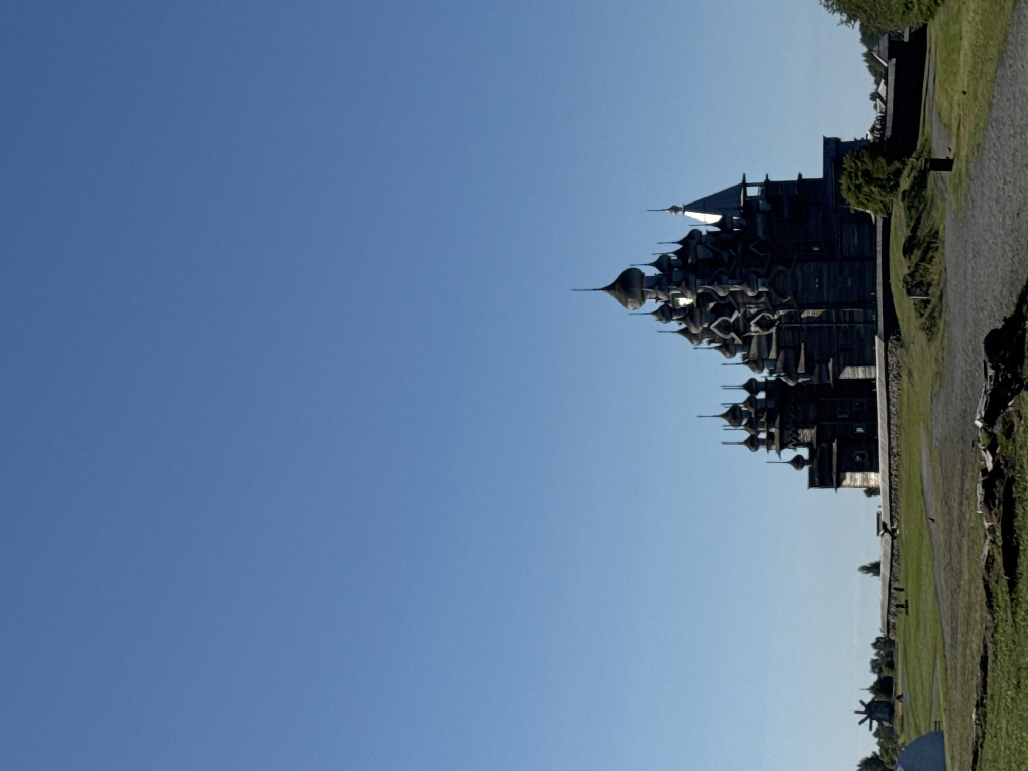 The image depicts the Kizhi Pogost, a UNESCO World Heritage site in Yamka, Russia, under a clear blue sky. The main structure is a complex wooden church with multiple domes and spires, appearing dark and aged. To the left of the church, a traditional wooden windmill stands on a grassy field. A stone wall encloses part of the area, with trees and other wooden structures visible in the distance. The foreground features a gravel path and green grass, with some rocks and debris scattered on the ground. The scene is an outdoor view of historical architecture in a rural setting.