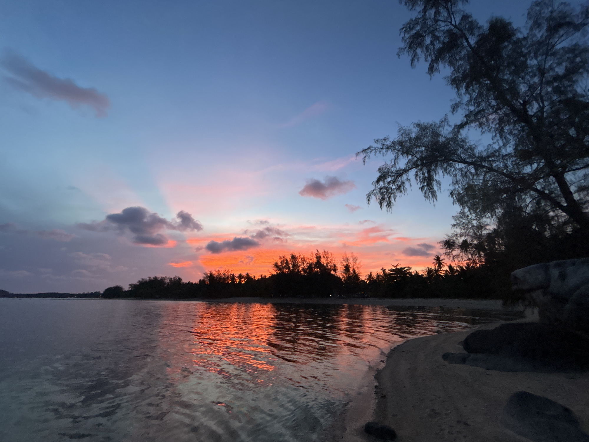 The image depicts a sunset over the ocean in Ban Bang Khao, Thailand. The sky is a gradient of colors, transitioning from blue at the top to pink and orange near the horizon. Clouds are scattered throughout the sky, some reflecting the colors of the sunset. The sun's rays are visible through the clouds. The water reflects the colors of the sky, creating a shimmering effect. In the distance, there is a line of trees silhouetted against the horizon. In the foreground, there is a sandy beach with some rocks. A tree is visible on the right side of the frame, partially obscuring the view.