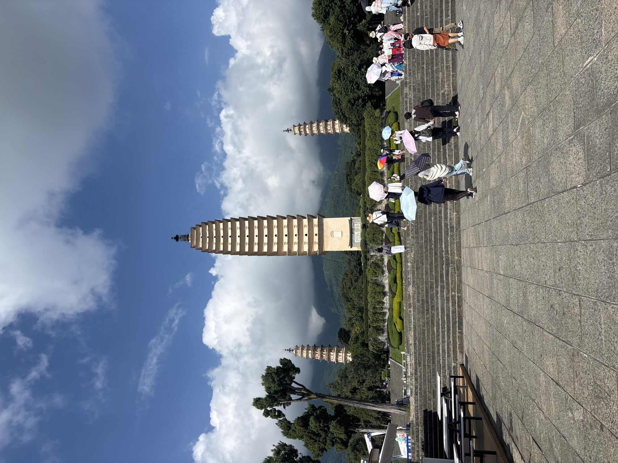 The image shows the Three Pagodas of Chongsheng Temple in Dali City, China. The pagodas are arranged in a triangular formation with the tallest pagoda in the center. They are surrounded by lush greenery and mountains in the background. Several tourists are visible in the foreground, many holding umbrellas, walking up a wide stone staircase. The sky is blue with scattered white clouds.