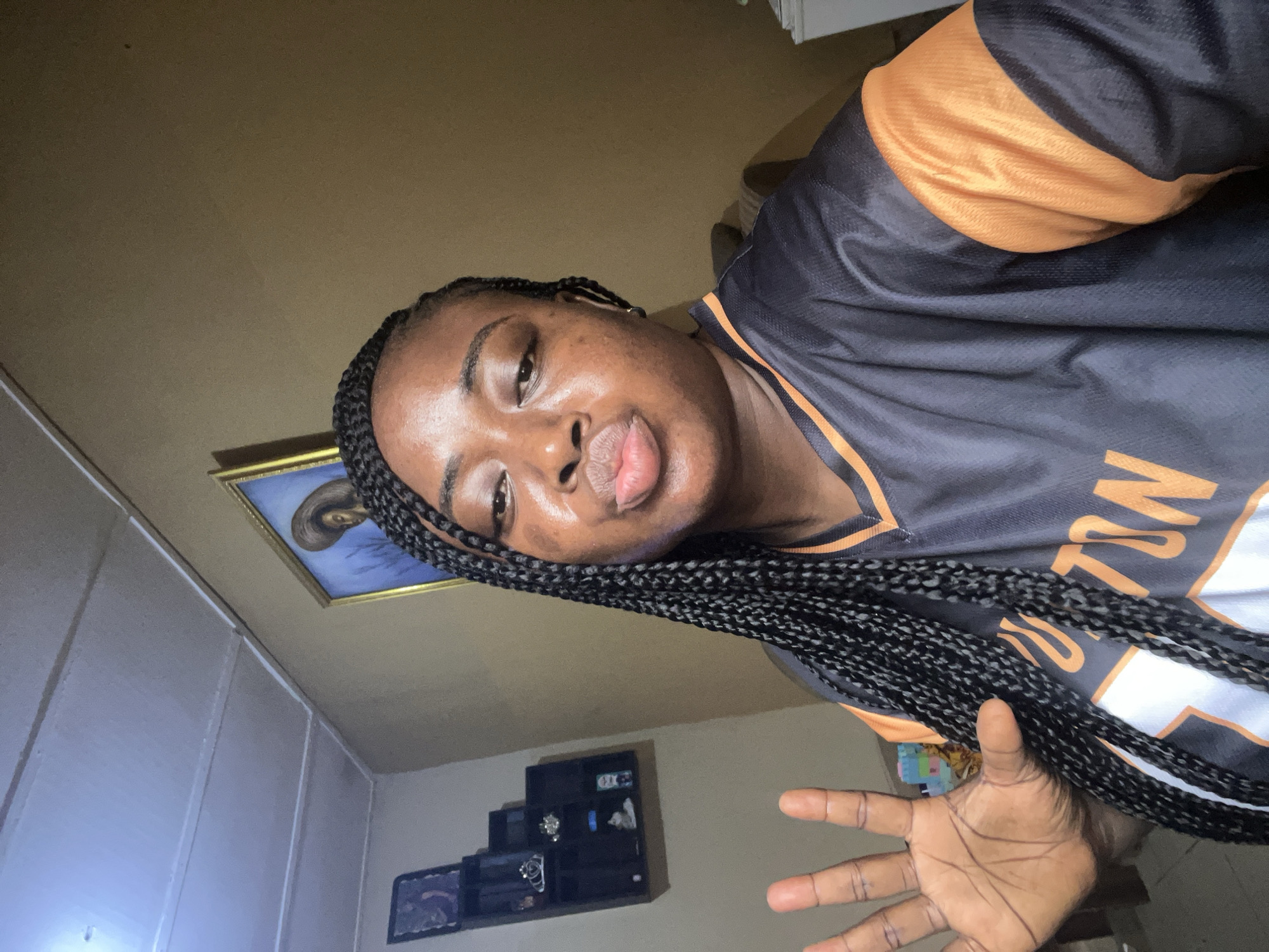 A photograph taken indoors, likely in a residential setting in Madalla, Nigeria, features a dark-skinned woman with braided hair, wearing a black and orange sports jersey. She appears to be holding up her hand. In the background, a picture frame hangs on the wall, and a small shelving unit displays various items. The room is dimly lit, and the walls are a neutral color.