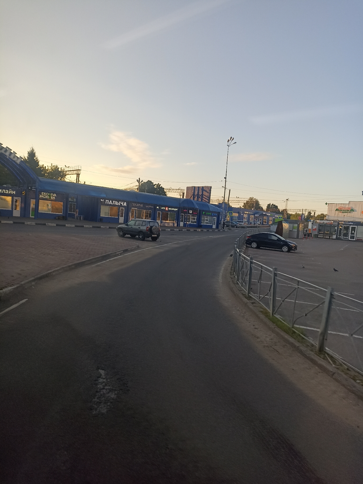 The image depicts a street scene in Chekhov, Russia, likely during the late afternoon or early evening, given the warm sky tones and the shadows. The foreground shows a two-lane asphalt road with some wear and tear visible on the surface. A curb separates the road from a pedestrian walkway and a parking area. Along the walkway, a row of blue commercial buildings with large windows lines the street. Signs on these buildings indicate various businesses, including "EKOPIVA," "ПАЛЬЧА," and "ПЕКАРНЯ."
Several cars are present. A dark-colored SUV is parked near the pedestrian walkway, and a black sedan is parked in the lot across the street. In the distance, more buildings and structures can be seen, including what appears to be a gas station or convenience store with a green sign. A tall lamppost with multiple lights stands near the commercial buildings. A metal fence runs along the perimeter of the parking area on the right side of the image. A few pigeons are scattered across the asphalt. The sky above is a soft blue with hints of orange and yellow near the horizon, suggesting a sunset or sunrise. Trees are visible behind the buildings on the left.