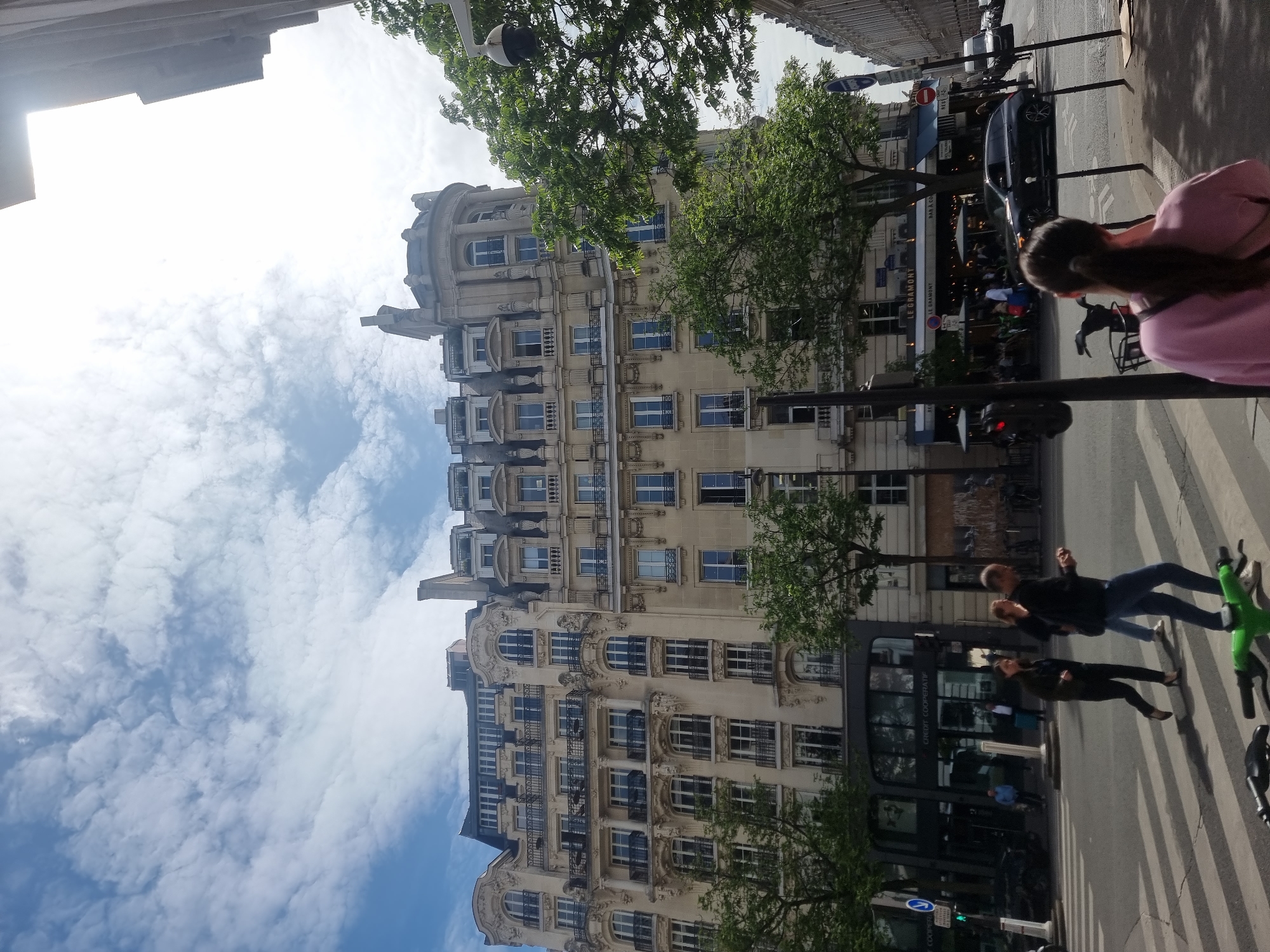 This is a daytime urban street scene in Paris, France, showing a bustling sidewalk and a grand, ornate building under a partly cloudy sky. The image is taken from a slightly low angle, looking up at the building and across the street.
The prominent subject is a multi-story building on the left, featuring classical architectural elements. It has a light-colored stone facade with numerous windows, balconies with intricate iron railings, decorative moldings, columns, and a distinctive domed top section. Trees with green foliage partially obscure the building.
On the right side of the street, several people are engaged in various activities. In the foreground, a person with brown hair in a pink top is seen from behind, walking toward the left. Further down, two individuals are crossing a street at a crosswalk; one man in a dark jacket and jeans is accompanied by a bright green electric scooter, while another person walks behind him. Further in the background, a café or restaurant named "LE GRAMONT" with an awning and outdoor seating is visible, where several patrons are seated. A black car is parked on the street near the café. Streetlights, traffic signs, and other street furniture are also present.
The sky is bright with scattered white cumulus clouds, indicating fair weather. Shadows suggest it is either late morning or early afternoon.
Visible text includes: "LE GRAMONT" and "BAR & CO" on the café awning, a red circular "SENS INTERDIT" (No Entry) sign, and "CRÉDIT COOPÉRATIF" on the facade of a building across the street, confirming a French location.