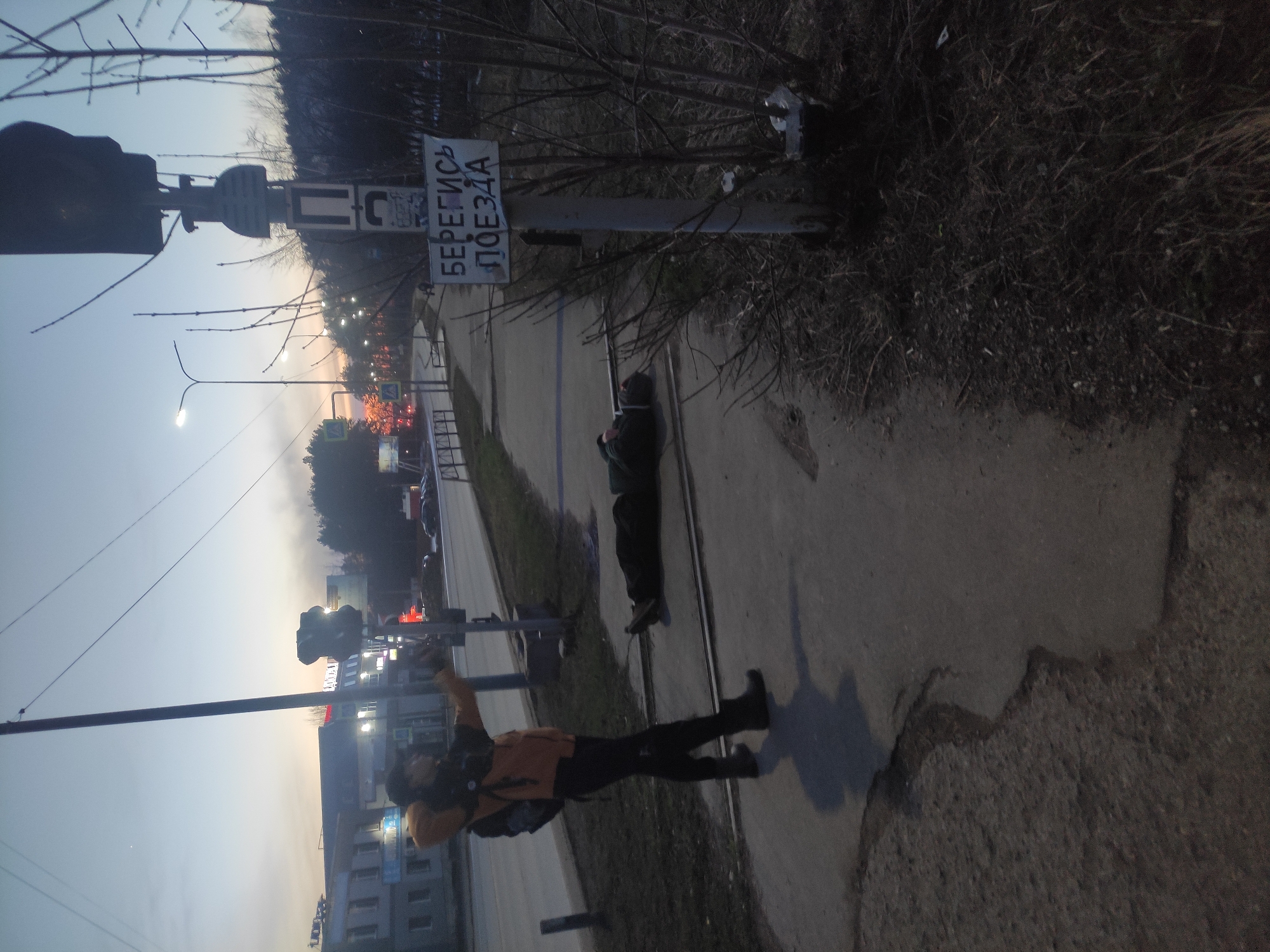 The image shows an urban scene in Chaikovsky, Russia. A sidewalk runs along a street with a tram line. A man wearing a dark jacket and pants, with an orange vest and a backpack, stands on the sidewalk, arms outstretched to either side. Nearby, another person is lying on the sidewalk, dressed in dark clothing. Adjacent to them is a sign that reads "БЕРЕГИСЬ ПОЕЗДА" (Beware of the Train). A railway crossing light post is visible next to the sign, and a traffic light is seen farther down the street. Streetlights illuminate the background, where buildings and trees are visible. The overall lighting suggests it is dusk or dawn.