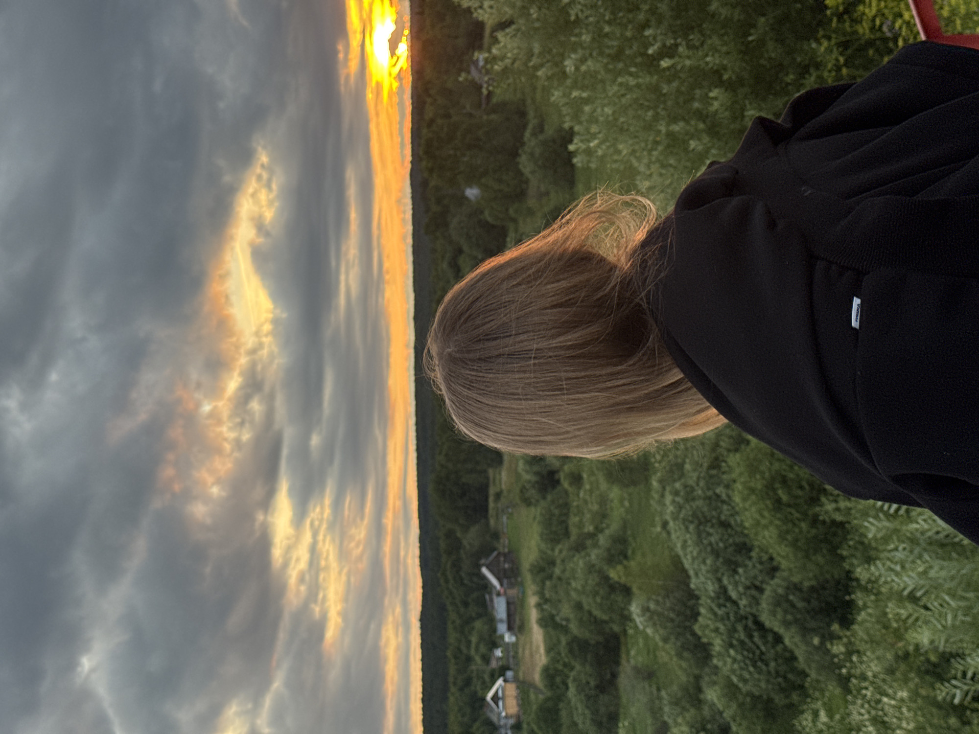 The image depicts a person viewed from behind, their head and shoulders in the foreground, looking out at a sunset over a rural landscape. The person has light brown hair and is wearing a dark hooded jacket. The landscape is characterized by rolling hills covered in dense green trees and some scattered houses and buildings in the distance. The sky is filled with dramatic clouds, with the sun setting in the distance, casting an orange and yellow glow. The overall impression is one of a peaceful, natural scene.