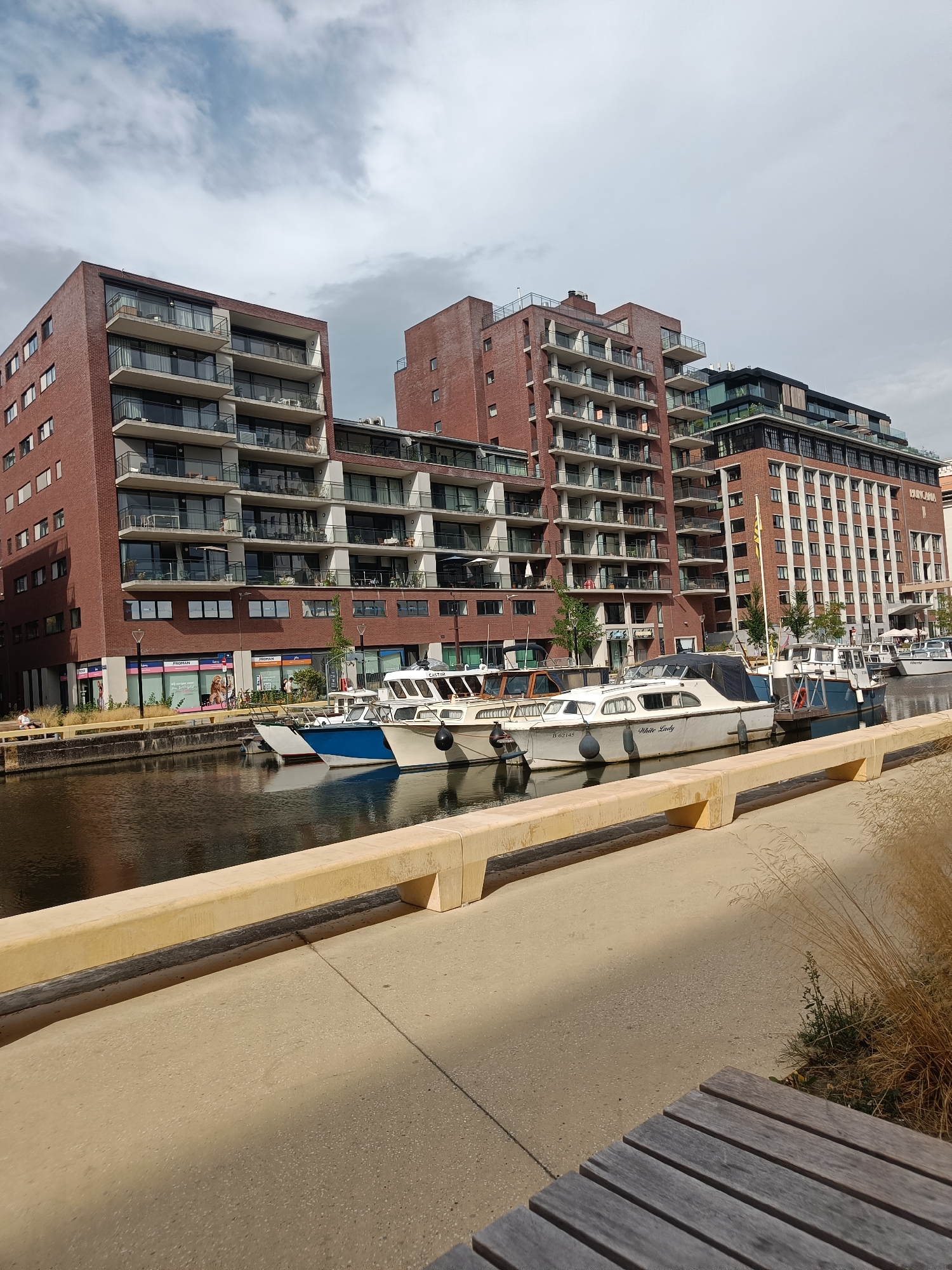 The image depicts a canal-side scene in Leuven, Belgium, likely near the Vaartkom area, characterized by modern architecture and leisure boats. In the foreground, a concrete walkway with a yellow concrete barrier runs alongside the canal. A wooden bench is partially visible in the lower right corner. The canal itself is dark and reflective, with several boats moored along the quay. These boats are of varying sizes and styles, with some appearing to be motor yachts.
Dominating the background are multi-story buildings constructed with red brick and large windows, interspersed with modern elements like balconies and glass facades. These structures suggest a mixed-use development, possibly residential and commercial. Signs on the buildings indicate businesses such as "PROMAN". The sky is cloudy, with patches of blue visible, suggesting a partially overcast day. A few trees are planted along the canal bank. There are no explicit signs of movement or activity from people in the immediate vicinity of the boats and buildings, though some figures might be present in the distant shopfronts. The overall impression is one of a contemporary urban waterfront.