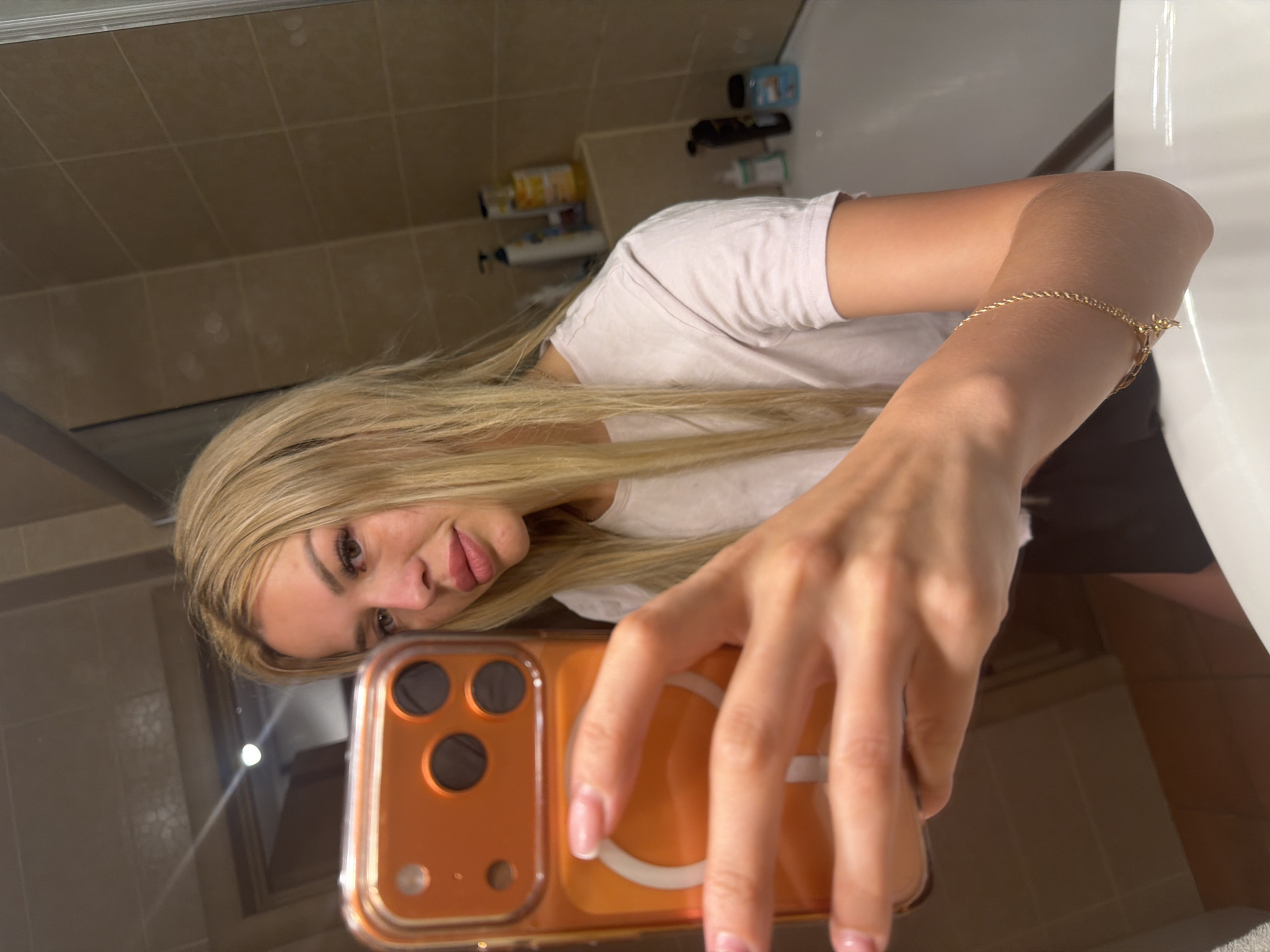 A young woman with blonde hair and full lips is taking a selfie in a bathroom. She is wearing a white t-shirt and has a gold bracelet on her right arm. She is holding an orange iPhone in her left hand. The bathroom has beige tiled walls and a white bathtub. There are several bottles of toiletries on a shelf. The image appears to have been taken in Kyiv, Ukraine, given the context of the request.