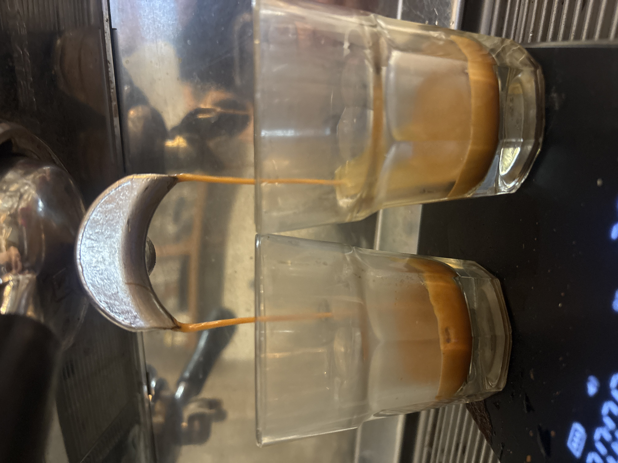 The image captures a close-up view of an espresso machine in the process of extracting two shots of coffee. Two thin, golden-brown streams of espresso pour simultaneously from the spouts of a metallic, dual-spout portafilter into two clear, small, rectangular glasses positioned below.
Each glass is partially filled with the beverage, showcasing its rich, caramel-like color and a visible layer of lighter, reddish-brown crema on top. The glasses themselves appear sturdy, with faceted sides near their bases.
The background reveals the shiny, metallic surfaces of the espresso machine, likely stainless steel or chrome, which reflect some ambient light. To the right of the glasses, a dark surface with faint blue light is discernible, possibly indicating a digital display or control panel of the machine. The setting appears to be an indoor environment, typical of a cafe or kitchen where coffee is prepared. No individuals are visible in the frame, nor are there any explicit textual elements, time-of-day indicators, or specific location cues within the image itself.