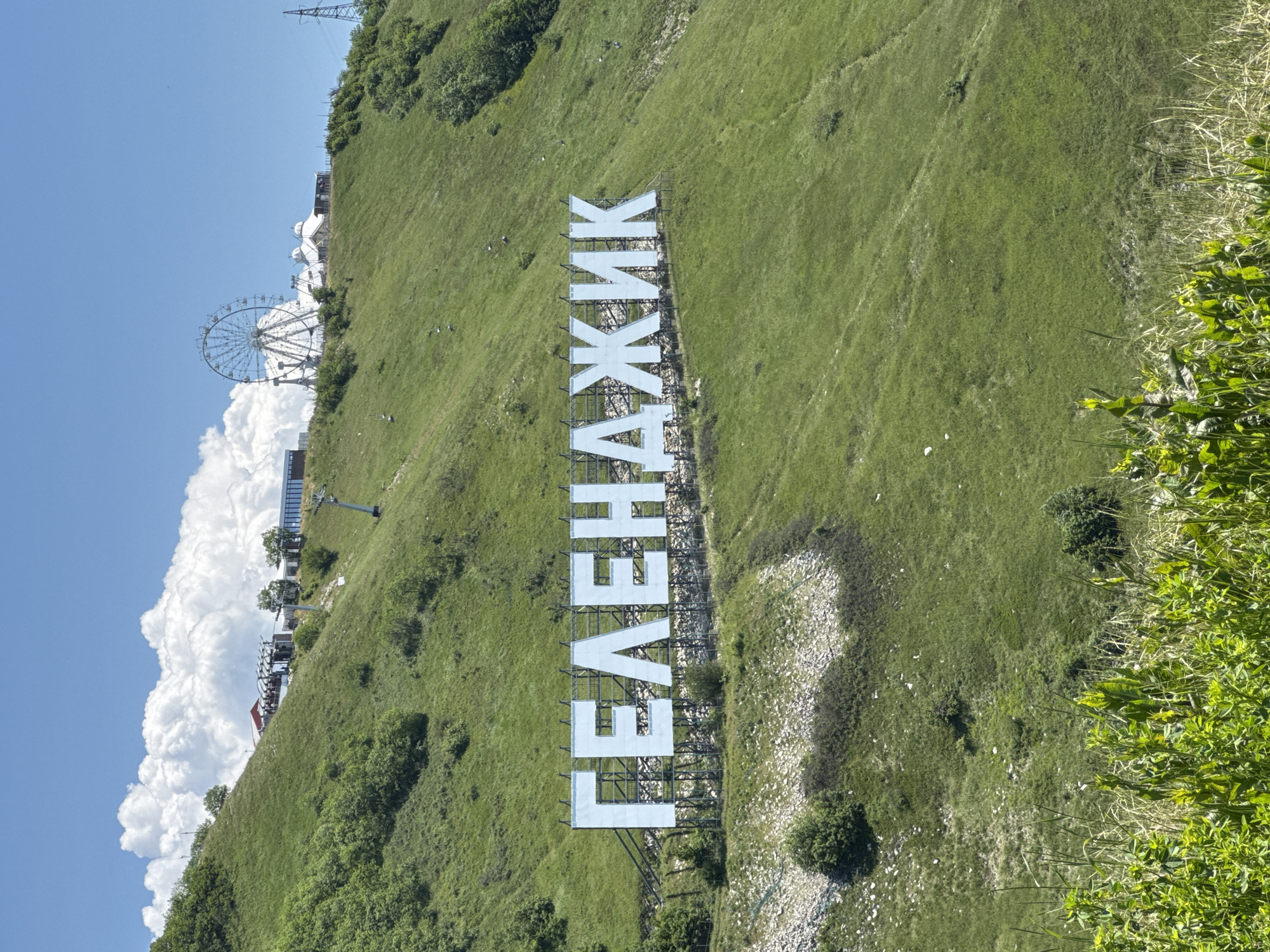 The image displays a prominent, large white sign spelling "ГЕЛЕНАДЖИК" (Gelendzhik) in Cyrillic, set against a verdant green hillside. The sign's letters are substantial, constructed from white panels supported by metal frameworks, evoking the style of the Hollywood sign.
The hillside itself is covered in lush green grass and scattered bushes, with some lighter, possibly rocky, areas breaking up the greenery. Above the sign, further up the slope and towards the left, a large white Ferris wheel is clearly visible, standing out against the bright blue sky. Adjacent to the Ferris wheel and along the hilltop, other buildings and what appear to be cable car or ski lift structures indicate an amusement park or tourist attraction at the summit.
The sky is a clear, vibrant blue, adorned with large, fluffy white cumulus clouds. The lighting suggests a bright, sunny day, likely mid-day, providing excellent visibility. The image captures a scenic view of a popular recreational area in Gelendzhik, Russia, blending natural landscape with man-made entertainment facilities.