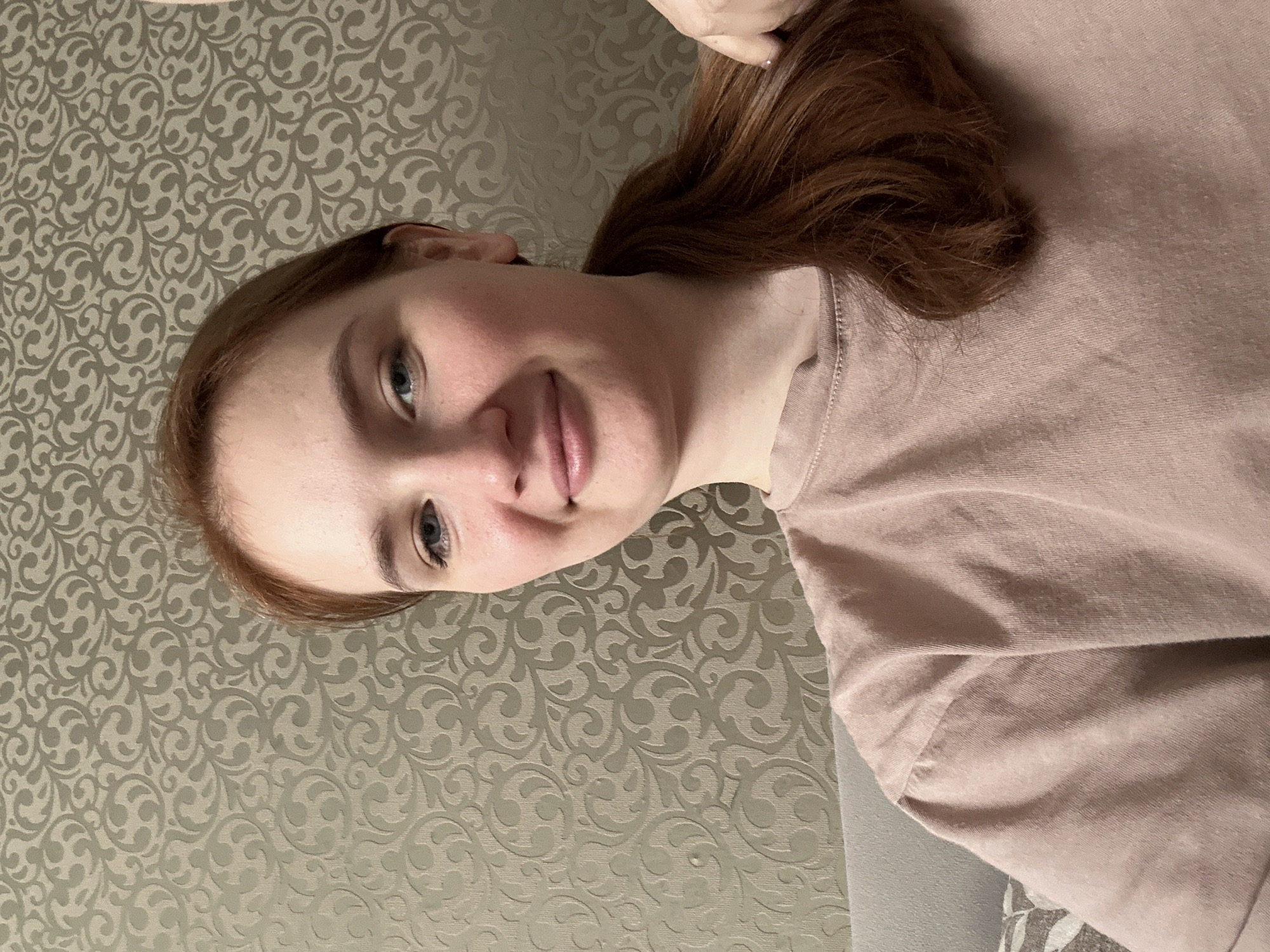 A woman with light skin and auburn hair is the subject of this close-up portrait. She is wearing a light pink T-shirt. The background features a patterned wallpaper with a repeating design of intertwined swirls and floral motifs in muted grey tones. The image appears to be taken indoors, possibly in a private residence. There is no evidence to suggest this image was taken in Chaikovsky, Russia. There is no indication of any actions or interactions taking place.