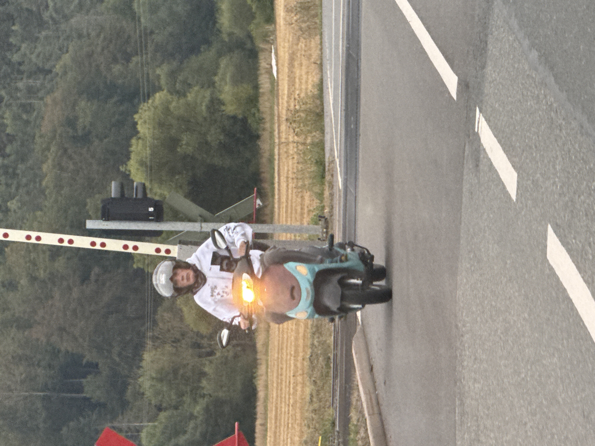 The image captures a person riding a blue and teal motor scooter through a railway crossing. The rider, wearing a helmet and a white hooded sweatshirt with a graphic design, is positioned centrally in the frame, approaching the viewer. The motor scooter's headlights are illuminated. In the background, a railway crossing gate with red and white markings and a traffic signal are visible. Beyond the crossing, a field of dry, light brown vegetation stretches towards a dense forest line. The scene appears to be outdoors, on a paved road with white lane markings. The presence of the railway crossing suggests a location near train tracks. The overall lighting and muted colors indicate it might be overcast or late in the day. The city of Ellrich, Germany, is not directly depicted in the image, but the context of a railway crossing is consistent with infrastructure found in such a town.