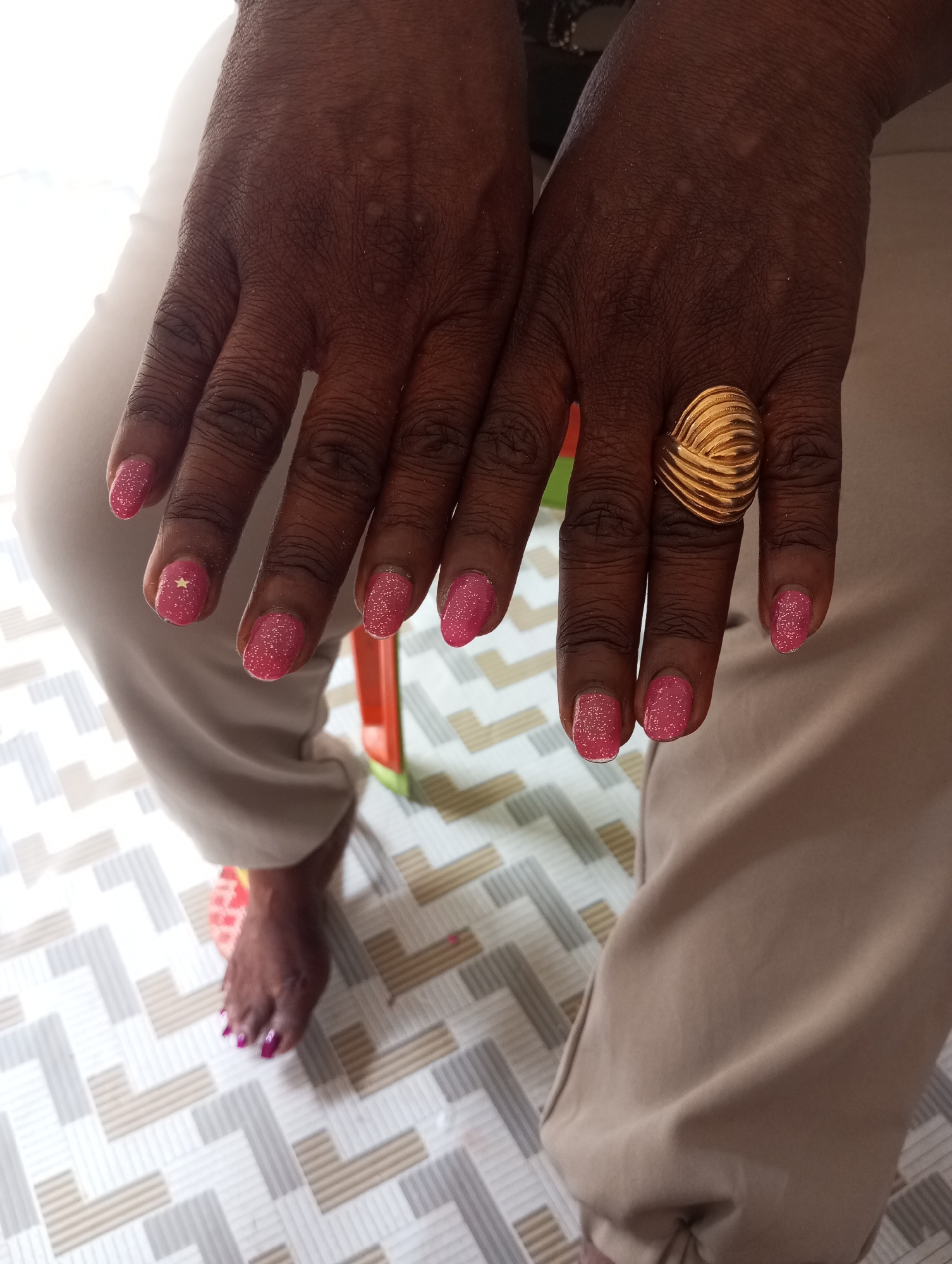 The image shows a person sitting with their hands and feet visible. The person is wearing beige pants and a gold ring on their right hand. The person's fingernails are painted pink with glitter, and one fingernail has a gold star sticker. The person's toenails are painted purple. The person is sitting on a chair with an orange back, and the floor is covered in a patterned tile. The location is Narayi, Nigeria.