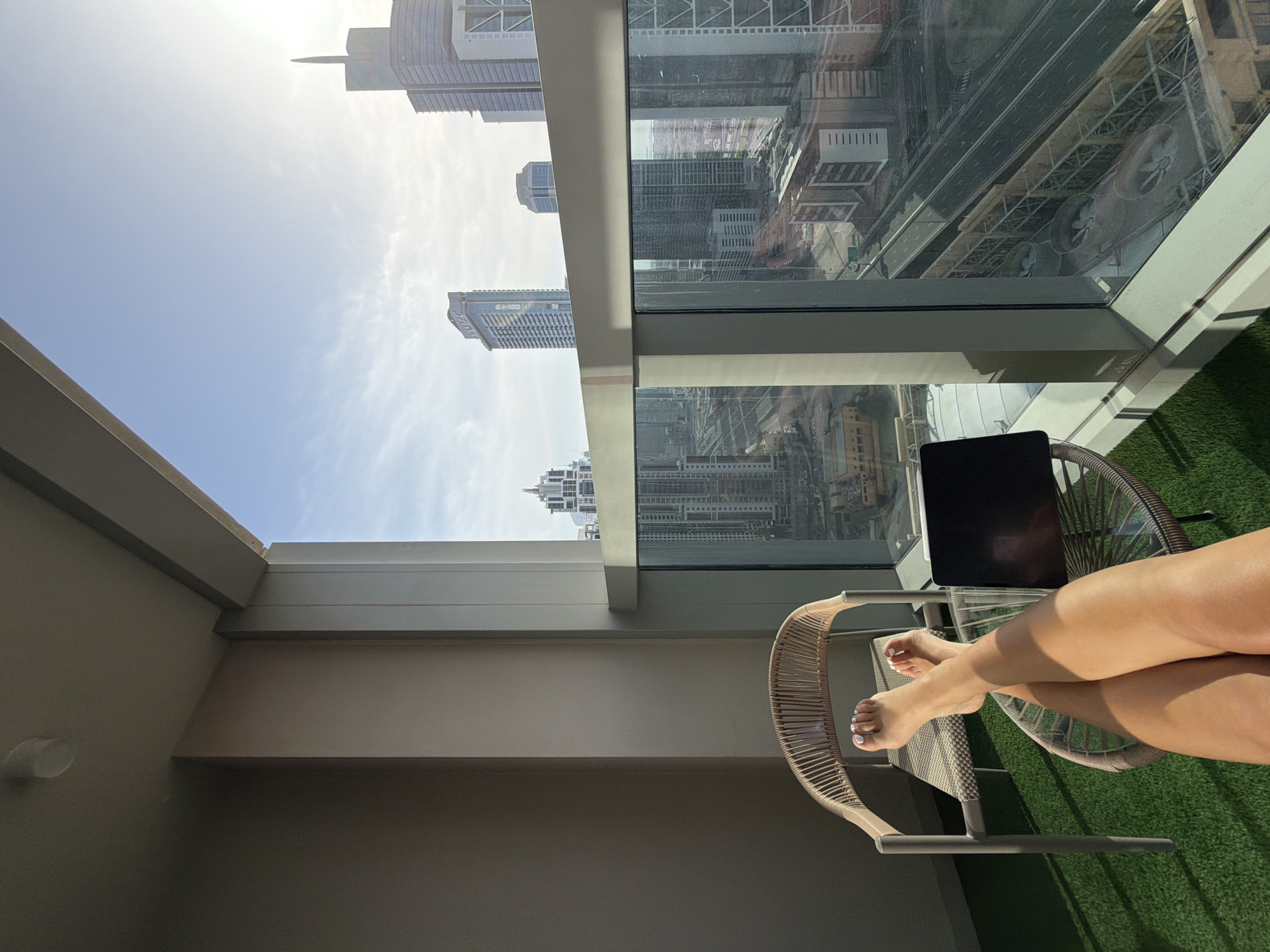 The image shows a view from a high-rise balcony in Business Bay, United Arab Emirates. The balcony features artificial turf. A person's legs are visible, crossed in a seated position in a wicker chair. A laptop rests on a glass table. Tall buildings are visible through a large window and beyond the balcony's open space. A bright, sunny sky is overhead. Reflected in the glass window are other city buildings, roads, and architectural structures.