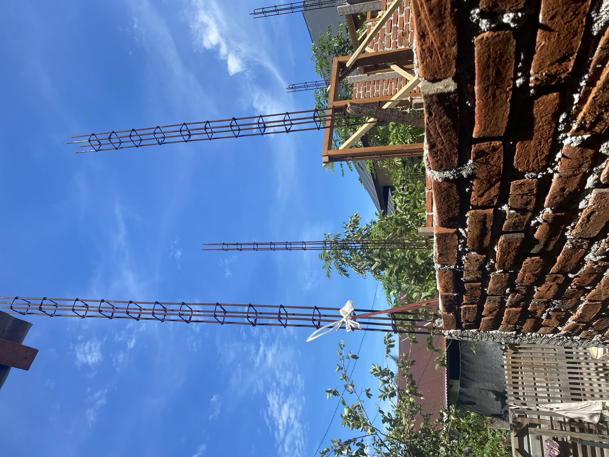 This image depicts an outdoor construction scene under a bright blue sky with scattered white clouds, likely in Banda Aceh, Indonesia. In the foreground, a brick wall, partially constructed, shows visible mortar between the red bricks. Behind the wall, tall, vertical rebar structures, designed to reinforce concrete columns, rise towards the sky. One of these rebar structures has a white plastic bag tied to it. In the midground, green foliage from trees and some reddish-brown corrugated metal roofing are visible, suggesting a residential or semi-urban environment. Wooden framing elements are also present, indicating ongoing construction work. The overall impression is one of a building in the early stages of construction.