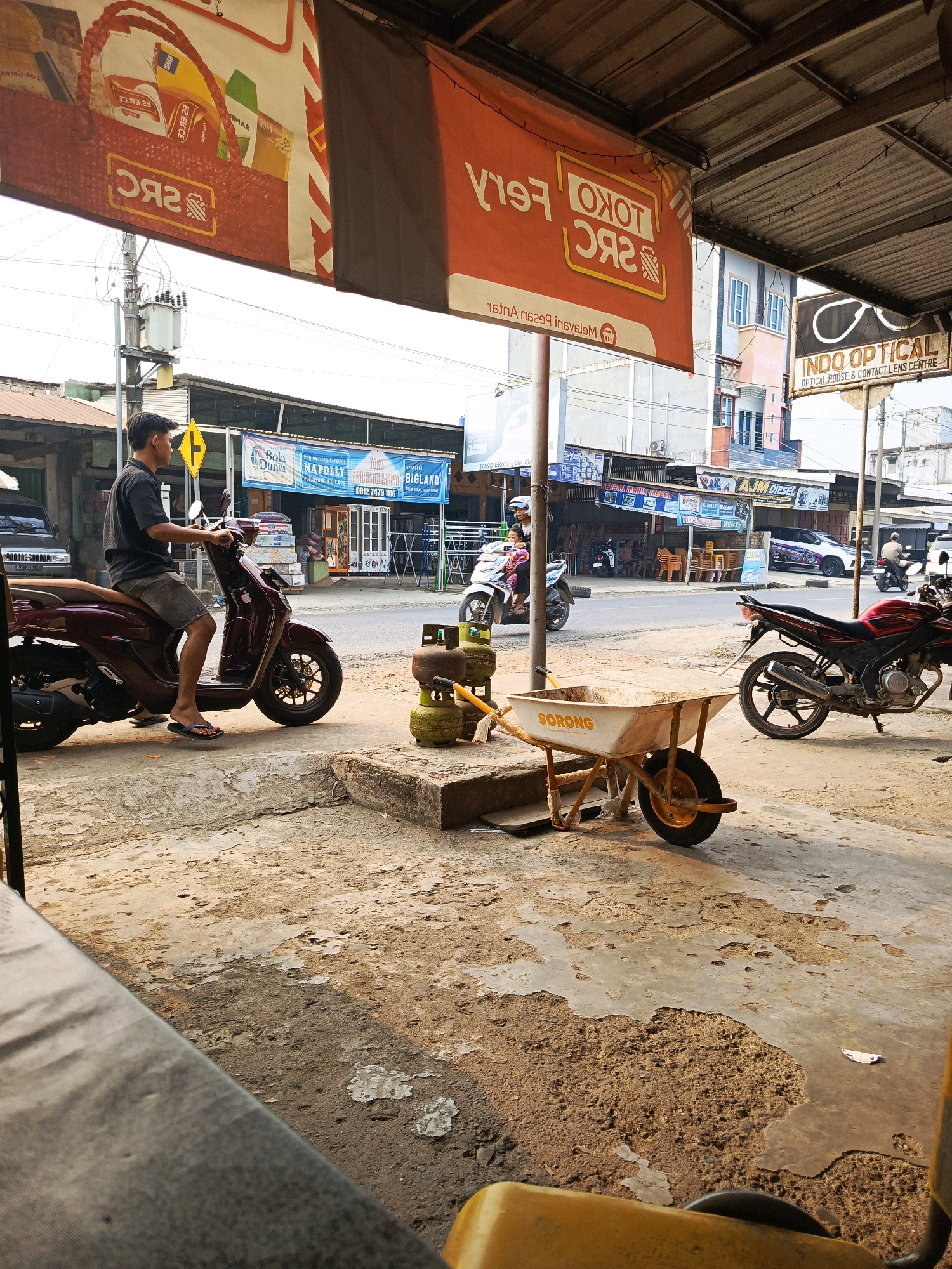 A man in a black shirt and grey shorts sits on a maroon scooter on a concrete sidewalk. He is facing right, looking ahead. Behind him, several green propane tanks are stacked on the ground. To the right of the tanks is a yellow wheelbarrow with the word "SORONG" printed on it. A red motorcycle is parked behind the wheelbarrow. In the background, across the street, are several buildings, including one with a sign that reads "INDO OPTICAL." The street is visible with a few cars and motorcycles. The sky is bright and the scene appears to be outdoors during the day, likely in a commercial area of Megang Sakti II, Indonesia. The overhead structure casts a shadow over the sidewalk.