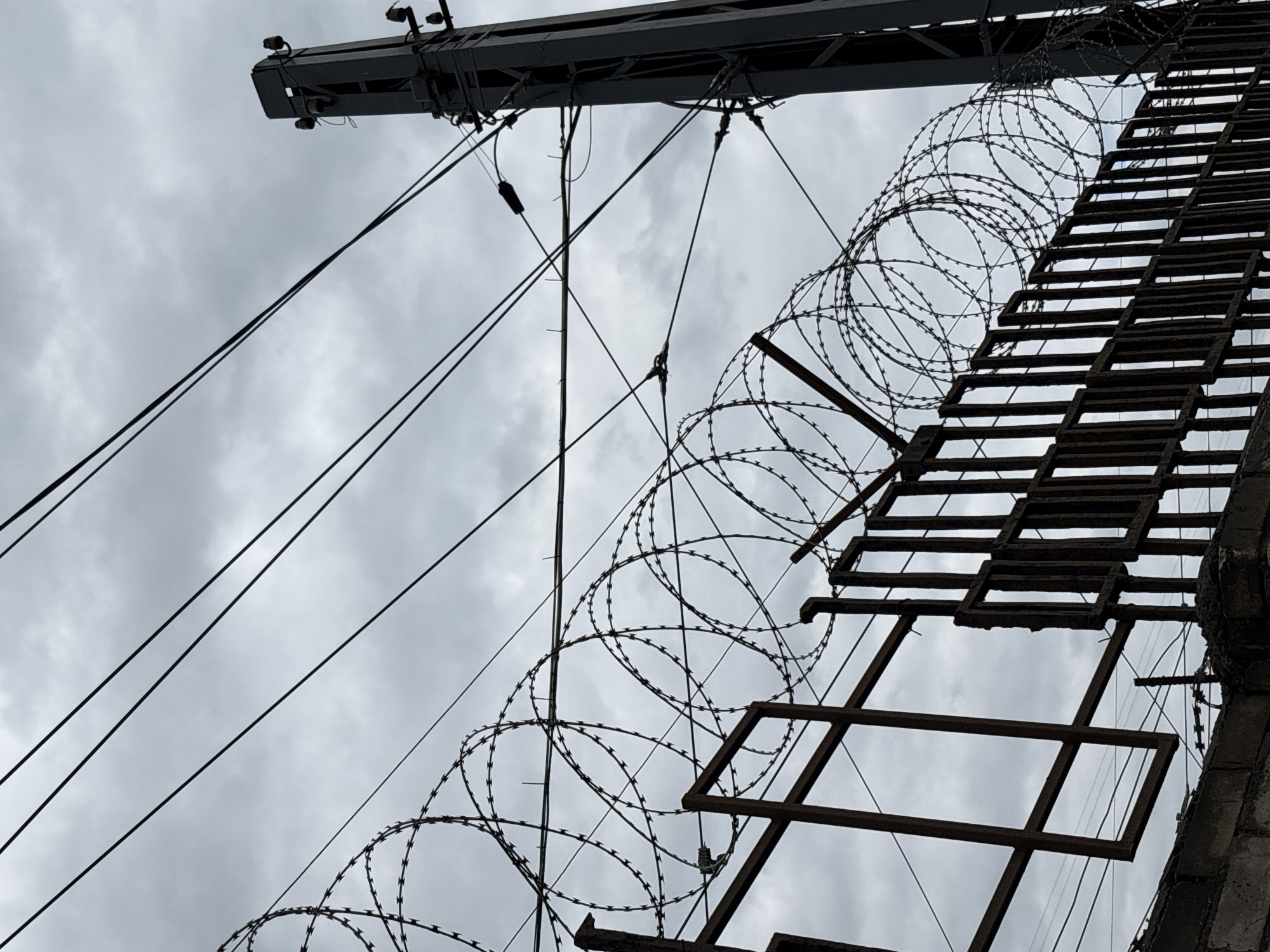 The image depicts a section of a prison facility. It features a ladder running up the side of a gray concrete wall topped with barbed wire. Above the ladder, there's a metal structure with lighting fixtures and numerous cables extending towards the sky, against an overcast, cloudy sky. There are no people visible in the frame. The location is Saint Petersburg, Russia.