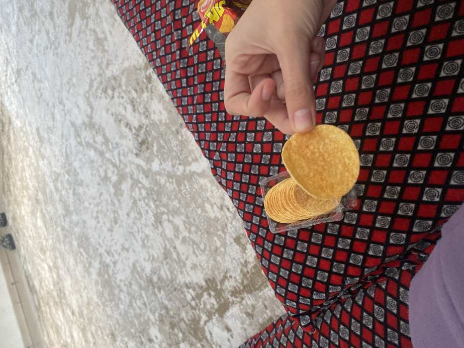 A person holds a round, golden potato chip above a small, clear plastic container filled with more of the same chips. The chips rest on a patterned surface of red, gray, and black diamonds containing rose-like shapes. A bag, likely holding more chips, sits slightly above the container. A light-colored, textured surface is visible to the left of the patterned surface. Part of a light purple sleeve is also visible.