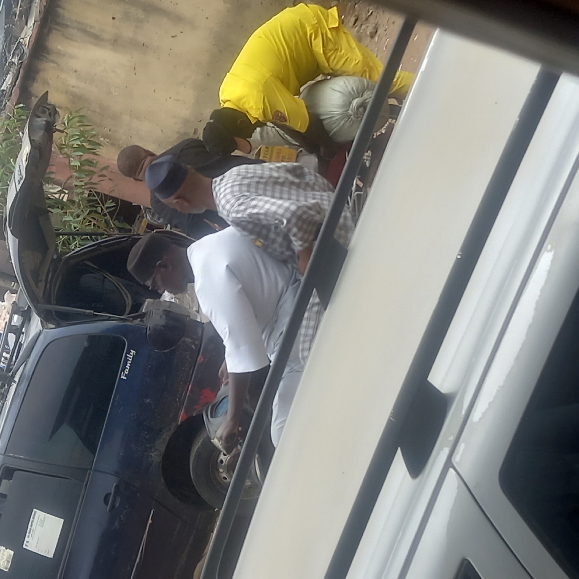 In Sardauna, Nigeria, the image depicts a group of people working on or near a dark blue van with the word "Family" visible on its side. One individual is dressed in a yellow outfit and is bending over. Another person is wearing a white outfit. The van has its hood open, and there is foliage visible nearby. A white vehicle is partially obscuring the view, and it appears the scene is viewed from inside the white vehicle.