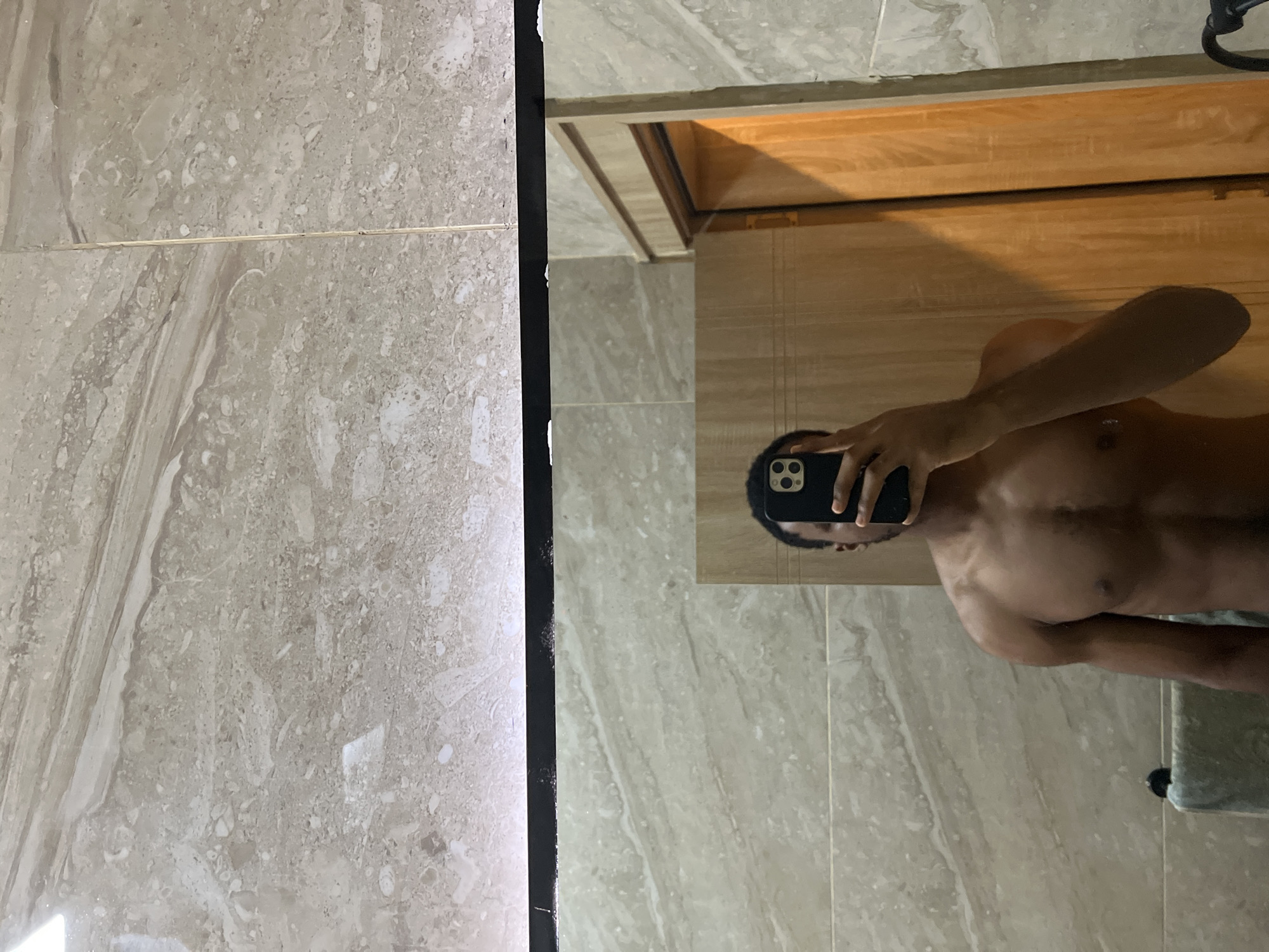 The image captures an indoor mirror selfie, likely taken in a bathroom or dressing area located in Gwarinpa, Nigeria. A shirtless person with dark skin is visible in the reflection, holding a dark-colored smartphone (featuring a distinct triple camera array) up to take the picture. Their face is obscured by the phone, but their torso, right arm, and part of their left arm are visible.
Reflected in the mirror are light grey and beige marble-patterned tiles, covering what appears to be the floor. Above the person's reflection, a wooden structure, possibly a cabinet or door frame, is also visible, casting a shadow that indicates an angled light source.
To the immediate left of the mirror, a wall is present, covered in similar light grey and beige marble-patterned tiles with visible grout lines. A thin, dark frame separates the mirror from this adjacent tiled wall. The entire scene is well-lit, highlighting the polished textures of the tiles and the person's skin. No discernible text is visible within the image.