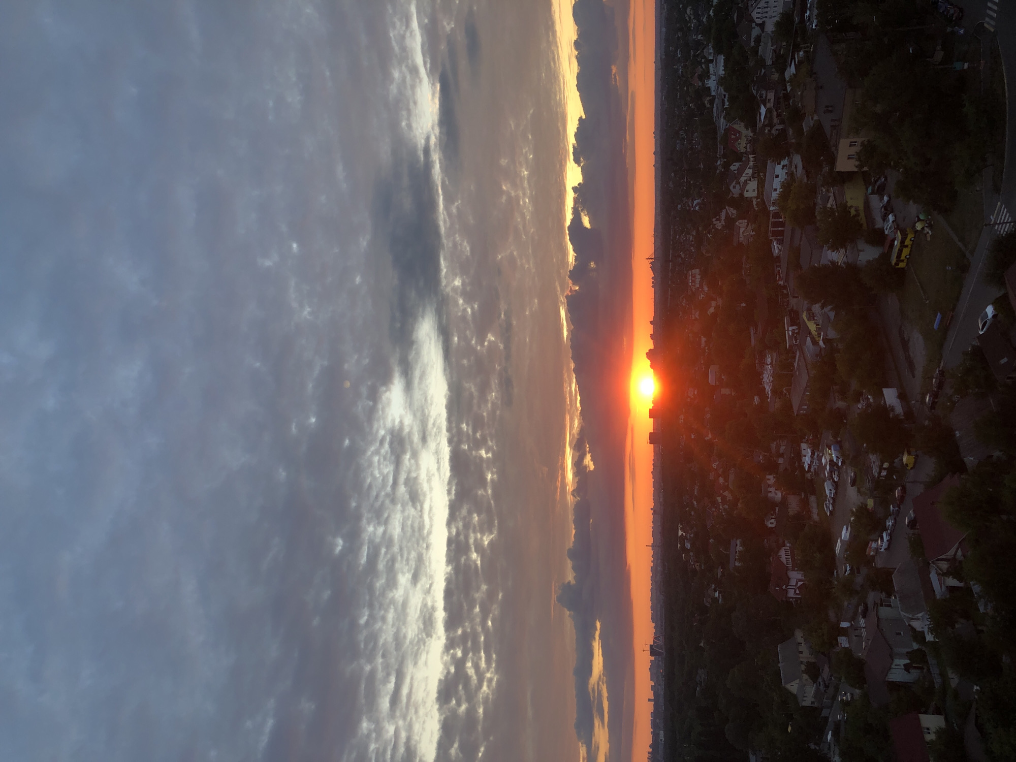 An aerial view captures a sunset over Kyiv, Ukraine, illuminating a sprawling residential area. The sun, a vibrant orange disc, is partially obscured by clouds, casting a warm glow across the sky and the landscape below. Residential buildings, interspersed with trees and streets, stretch towards the horizon, where the faint outlines of the city's skyline are visible. The sky is a dramatic canvas of oranges, reds, and purples, transitioning into cooler blue and gray tones higher up, with scattered clouds adding texture to the scene.