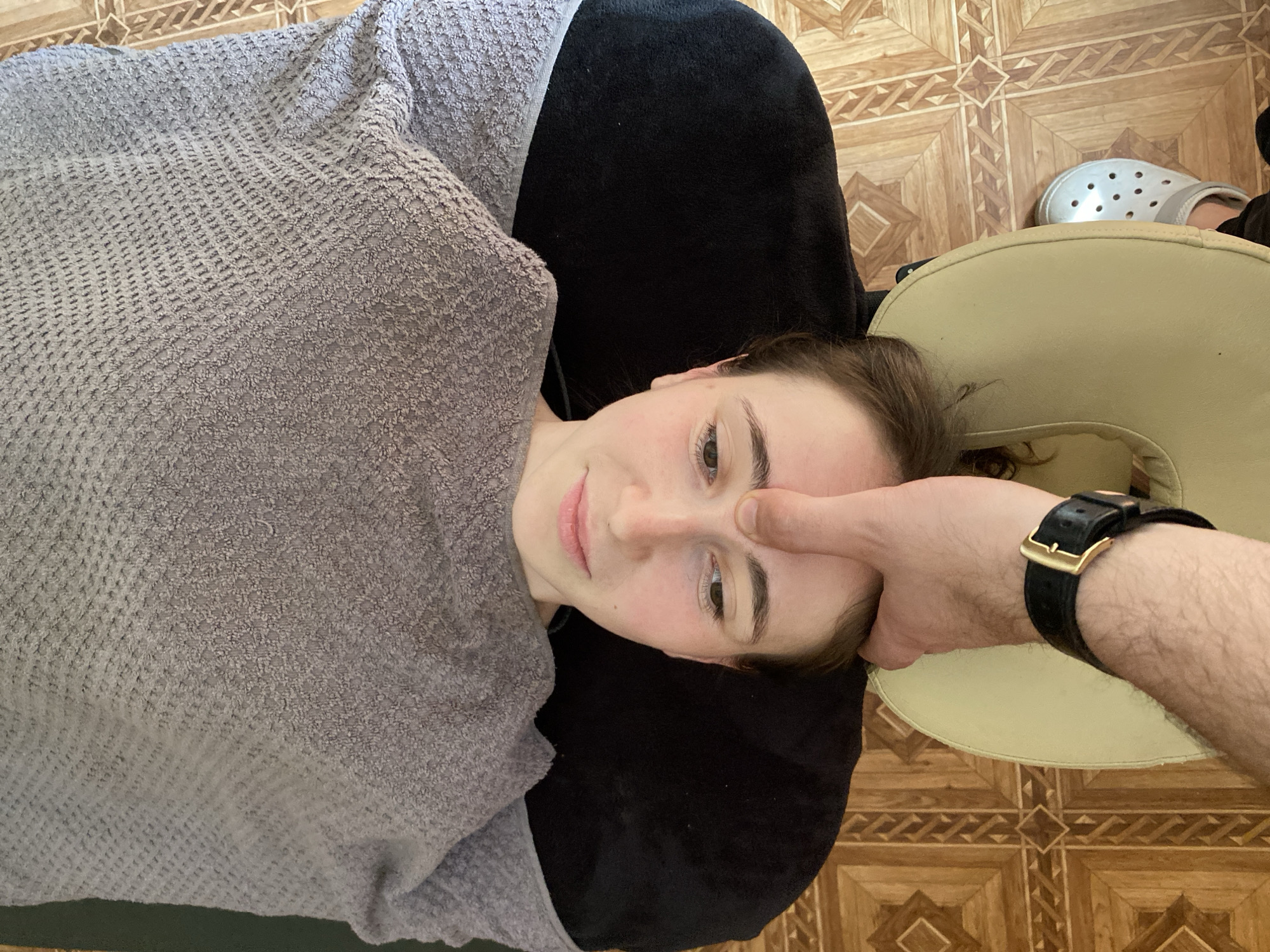 The image shows a top-down view of a young woman lying down, her head resting on a black pillow. She is covered by a grey knitted blanket. A person's hand, wearing a black leather watch with a gold buckle, is gently pressing on her forehead. The woman has light skin, brown hair, and is looking upwards with a neutral expression. Her eyes are open, and her lips are slightly parted.
The setting appears to be indoors, with a tiled floor visible in the background. The tiles have a wooden pattern with a geometric design. A light-colored massage face cradle is also visible. The lighting suggests it is daytime.
The image seems to capture a moment during a massage or similar therapeutic session, given the position of the subjects and the presence of the face cradle. There is no visible text in the image.