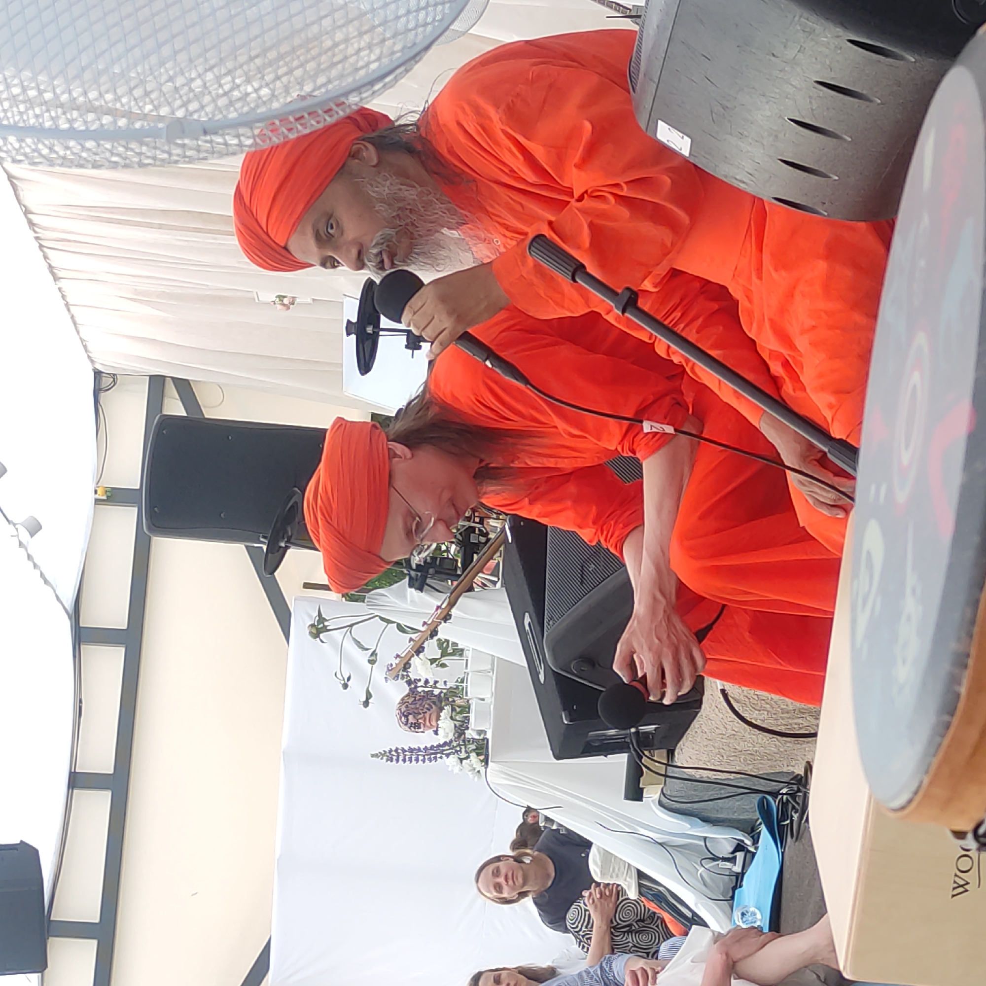 In Zhirnosovo, Russia, two men in orange robes and turbans are seated on the ground, facing right. The man on the left wears glasses and has long brown hair. He is looking down and appears to be speaking into a microphone, held by an unseen person to his right. The man on the right has a grey beard and is speaking into a microphone. Both are positioned in front of large black speakers. In the background, white drapes are visible, suggesting an outdoor or tented event. Several people are seated in the background on the left side of the image. The scene appears to be a performance or presentation.
