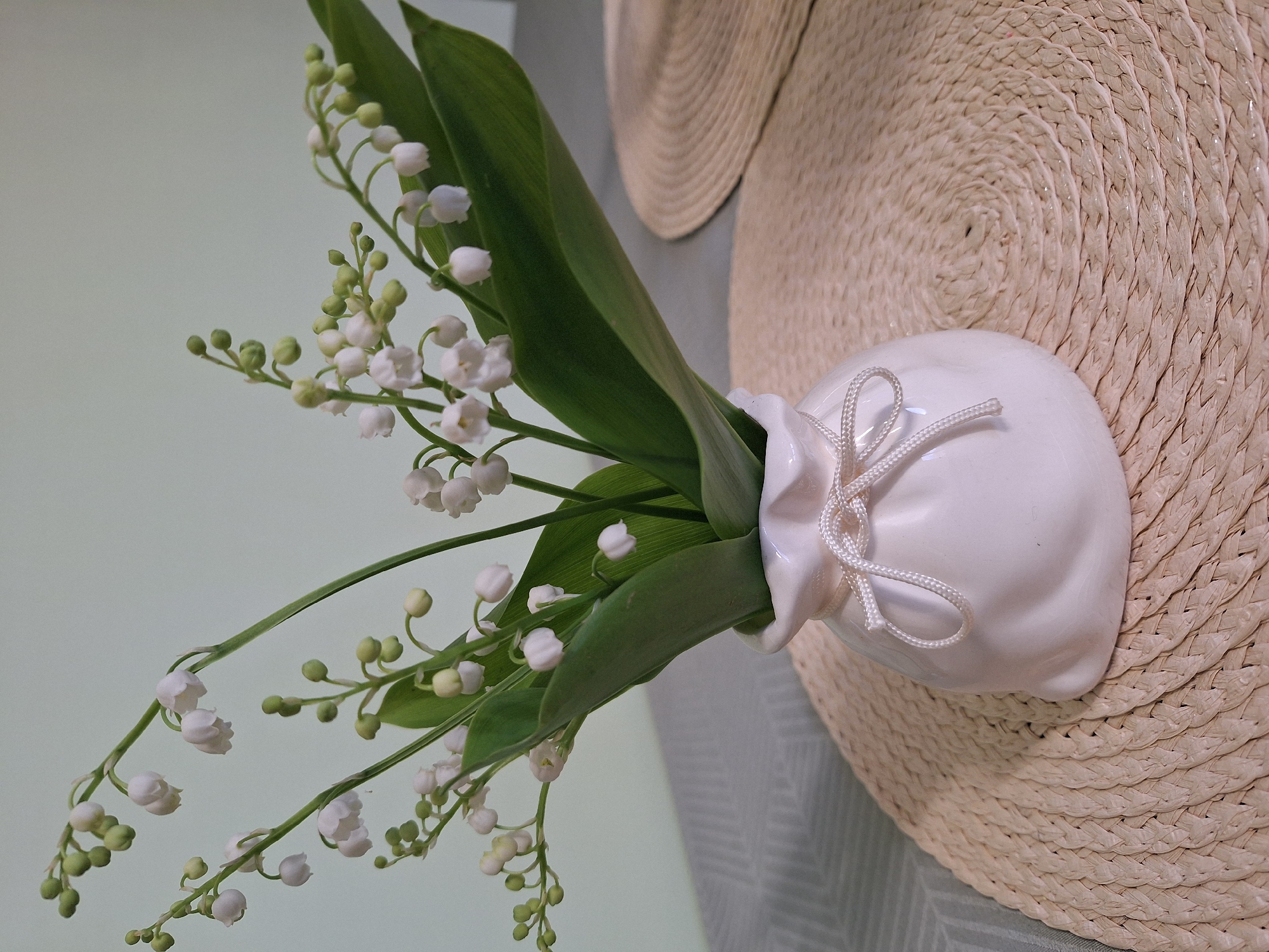 The image shows a white ceramic vase containing lily of the valley flowers with green leaves and stems. The vase has a string tied around its neck. The vase sits on a straw hat. The background is a light green wall and a grey patterned surface.