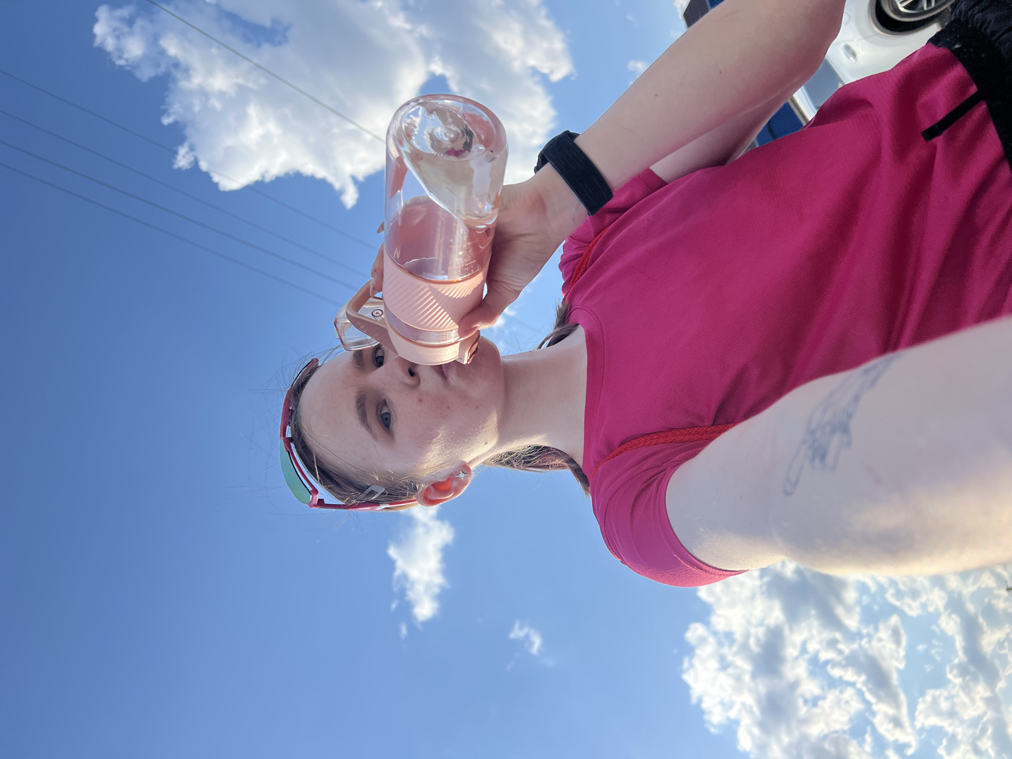 A woman is taking a selfie while drinking from a pink water bottle. She is wearing a pink shirt and a black fitness tracker on her wrist. The background is a blue sky with scattered white clouds. Power lines are visible in the upper left corner. The location is unknown, and there is no indication that the image was taken in Perm, Russia.