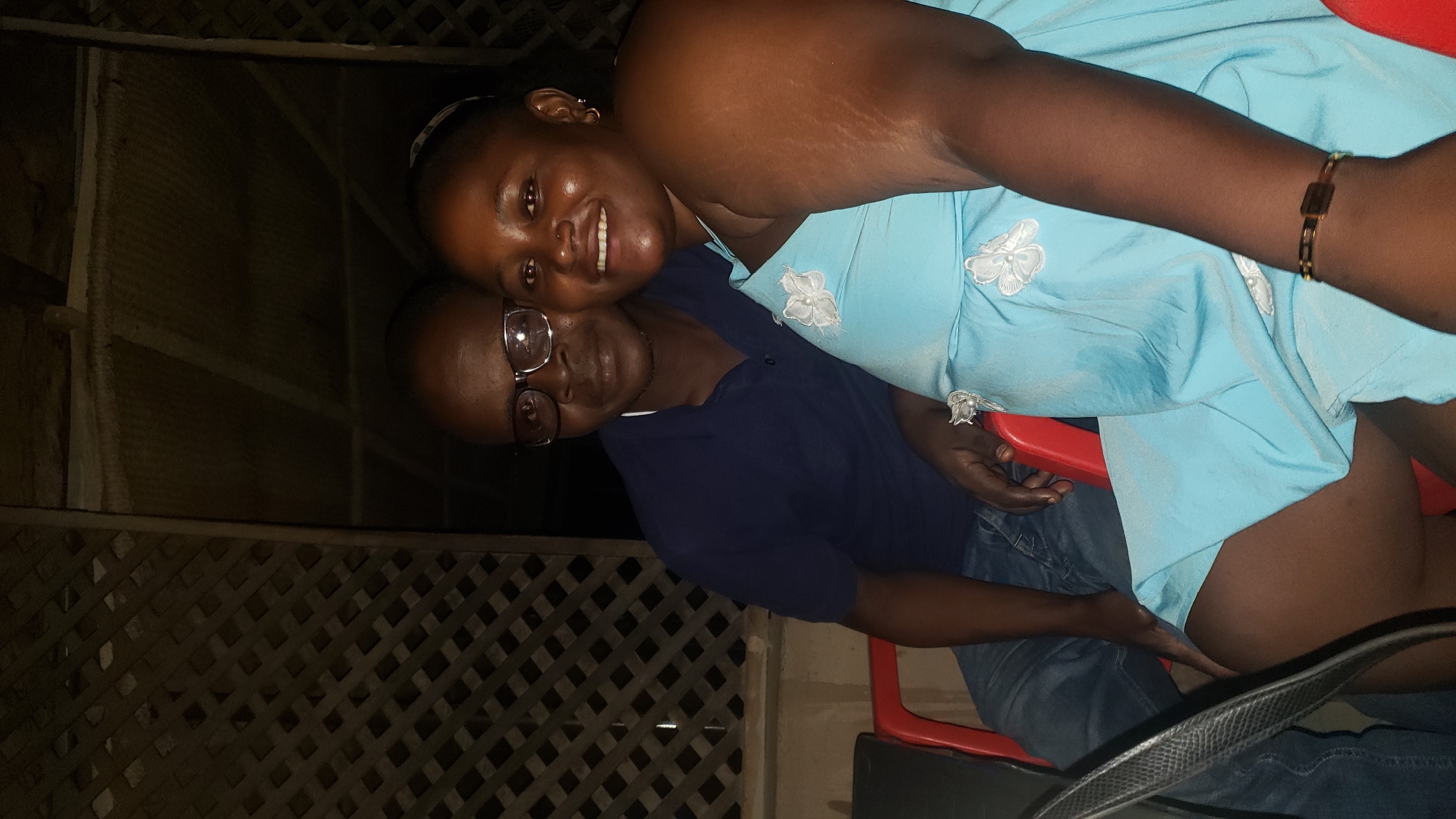 Two adults are posing for a photo in what appears to be an outdoor setting. A woman with a light blue dress and a bracelet on her right wrist is smiling. Adjacent to her is a man wearing glasses and a dark blue shirt. They appear to be sitting on red chairs. A lattice structure is visible in the background, possibly part of a building or fence. The location may be Narayi, Nigeria.