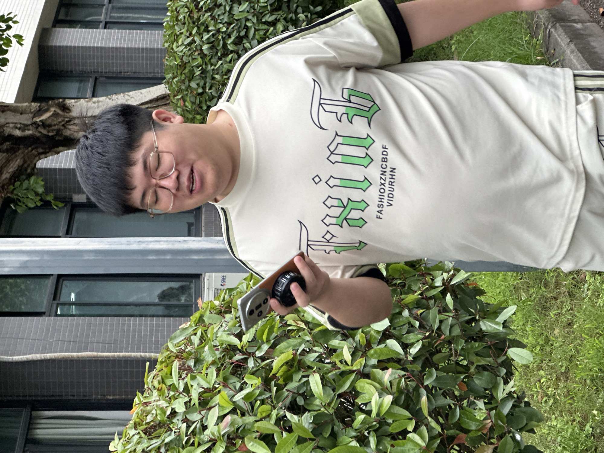 A man is depicted outdoors, holding a mobile phone and looking down at it with his mouth slightly ajar, suggesting he might be speaking or engrossed. He is wearing clear-framed glasses and a light-colored (cream or off-white) short-sleeved t-shirt with black and light green stripes on the sleeves. The front of his shirt features large, stylized green text, accompanied by smaller black text reading "FASHIOXZNCBDF VIDURHN". The mobile phone in his right hand has a black, circular accessory attached to its back, possibly a grip or a camera lens.
The background features dense green foliage or bushes in the mid-ground and foreground. Behind the man and the bushes, a building is visible, characterized by dark-framed windows and a grey tiled or brick-like facade. An outdoor unit with the brand name "GREE" printed on it is attached to the building. A tree trunk is partially visible on the left side, and a patch of green grass can be seen to the right of the bushes. The scene is brightly lit, indicating it is daytime. The image is oriented with the man appearing to lie horizontally, implying a 90-degree clockwise rotation from its intended upright view.