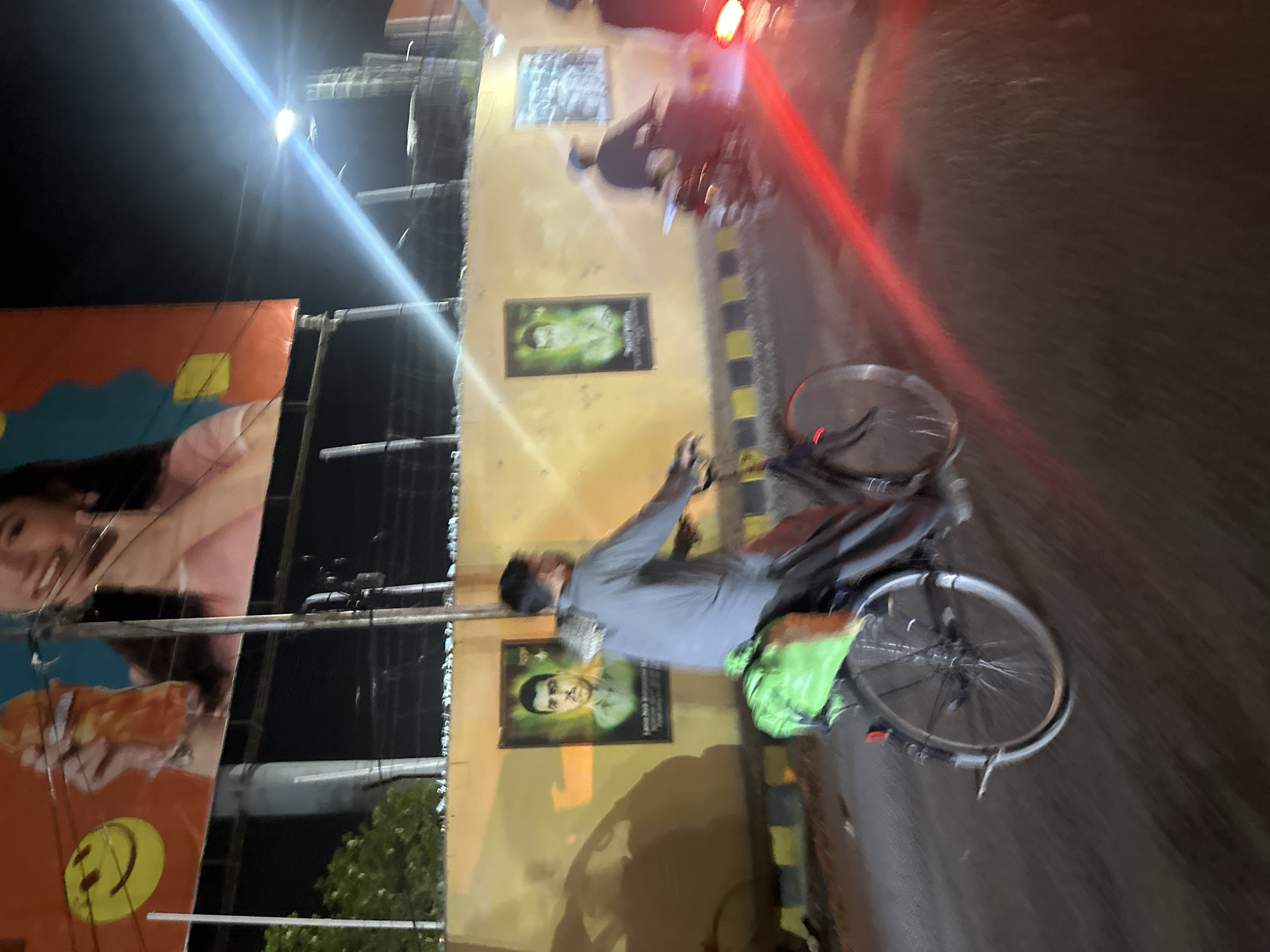 A bicyclist is riding on a paved road adjacent to a wall. The location appears to be Quetta City Tehsil, Pakistan. The wall is decorated with portraits. There's a large advertising billboard on the left. Street lights are visible. A motorcycle is present in the background. The time is likely night. The cyclist is wearing a grey shirt and is moving forward.